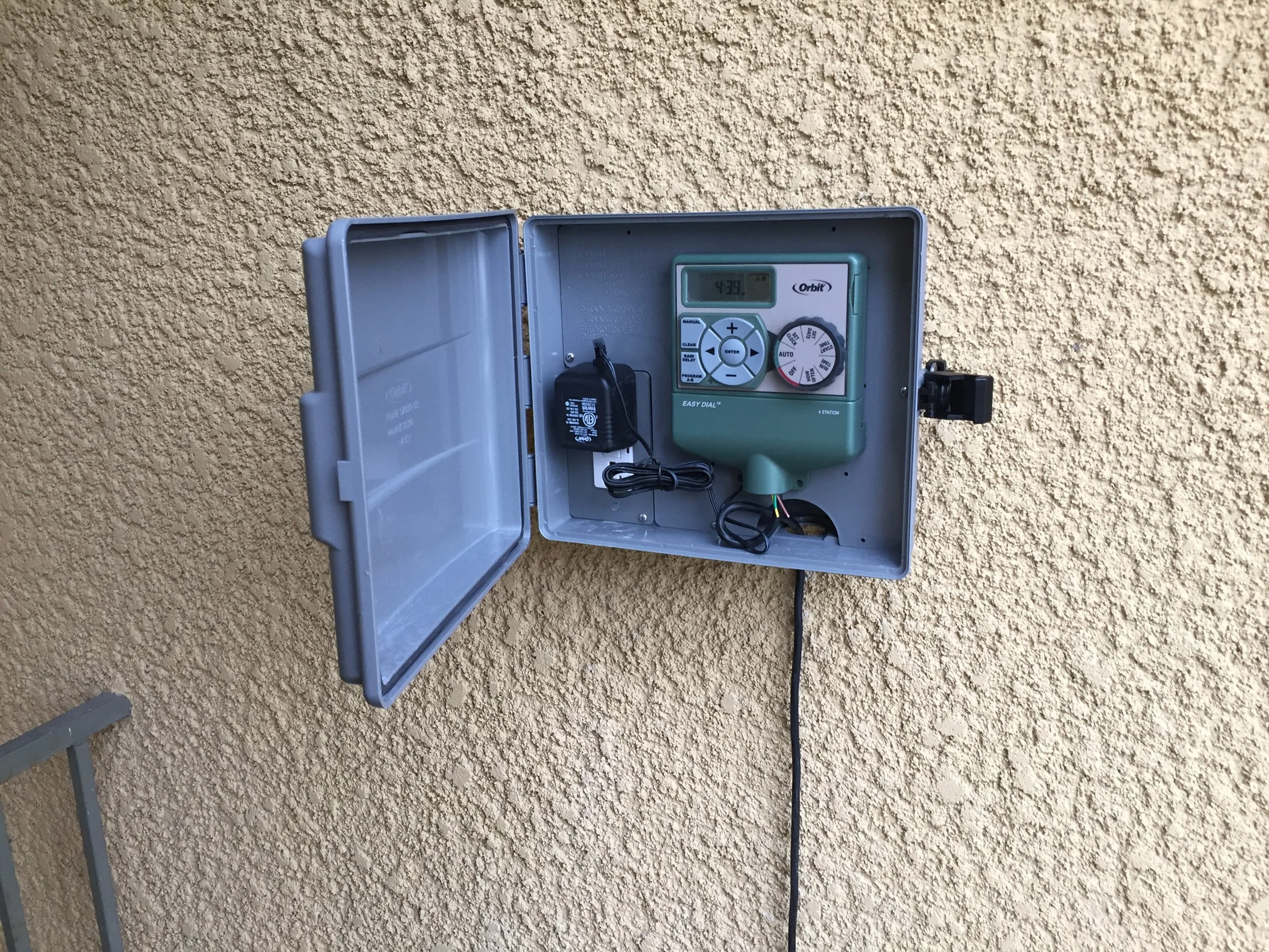 A gray outdoor electrical box mounted on a yellow stucco wall containing a green timer control device and a black power adapter.