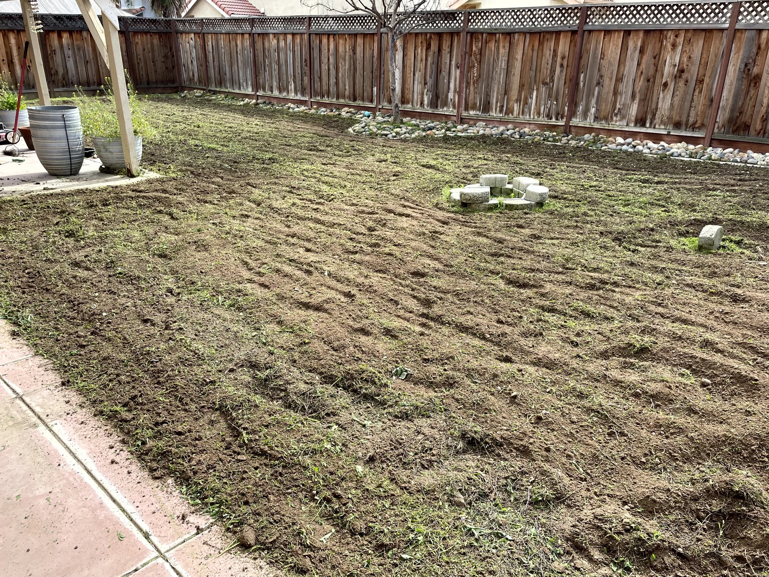 A backyard with freshly tilled soil, a wooden fence, potted plants, and a small circle of cinder blocks arranged for a fire pit.