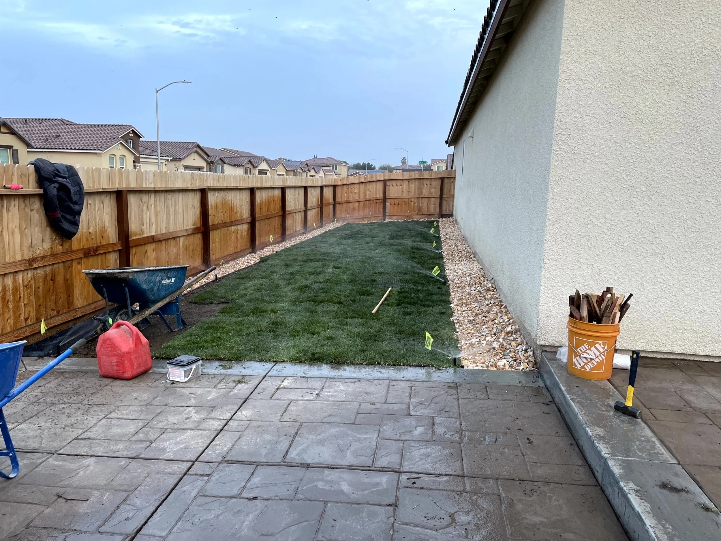 Backyard with a new lawn being installed, construction tools and materials visible, including a wheelbarrow, a red gas can, and a bucket of wood, with a wooden fence and houses in the background.