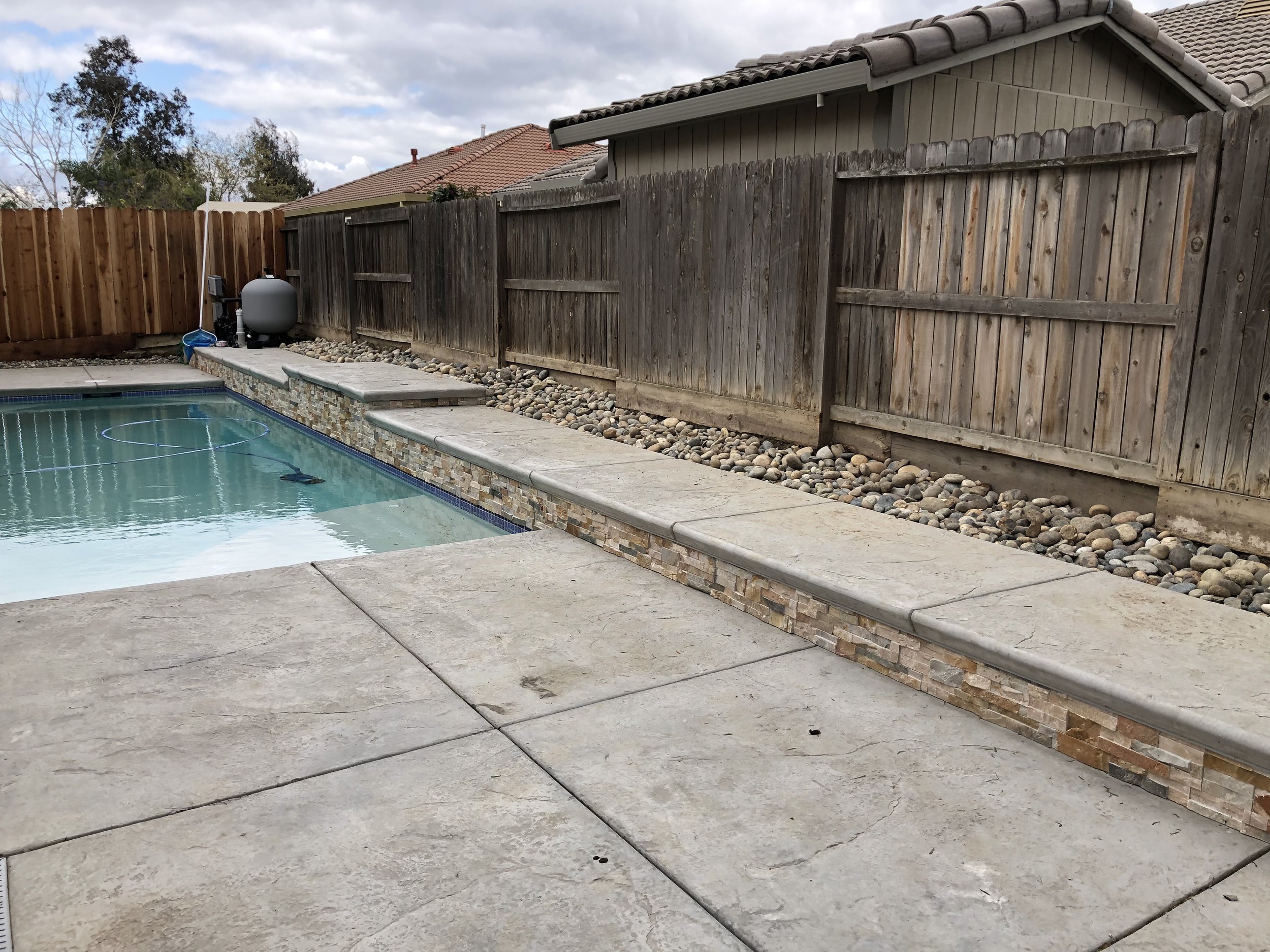 Backyard swimming pool with a stone fountain feature along the edge, surrounded by concrete patio and wood fencing.