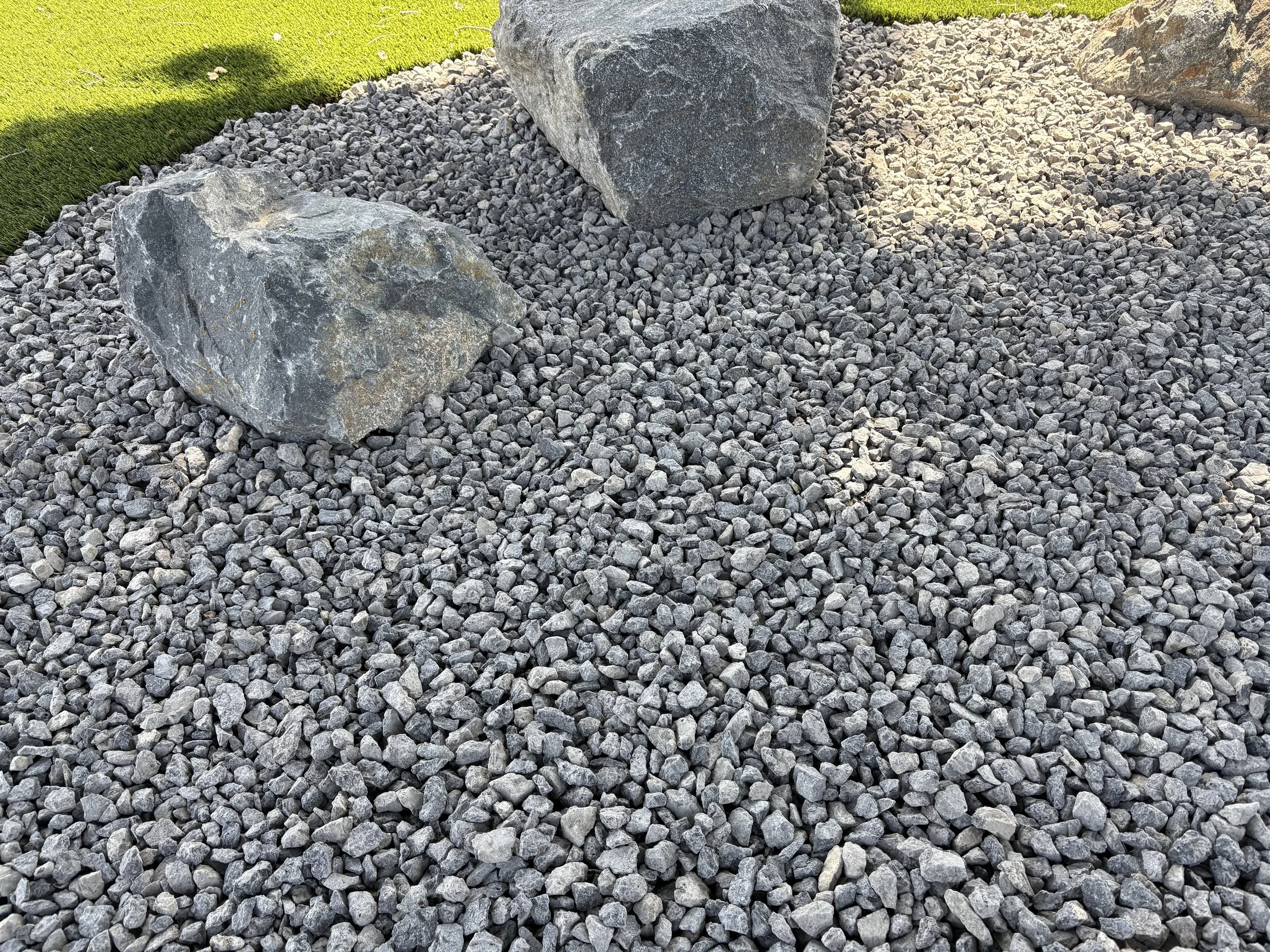 A garden landscape featuring large decorative rocks surrounded by small gray gravel, with a well-maintained grassy area in the background.