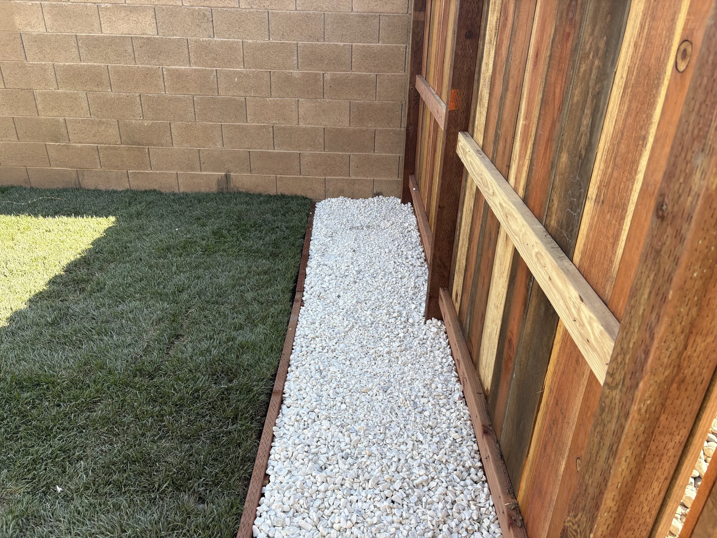 A backyard with green grass, a beige brick wall, a wooden fence on the right, and white gravel in between, bordered by wooden planks.