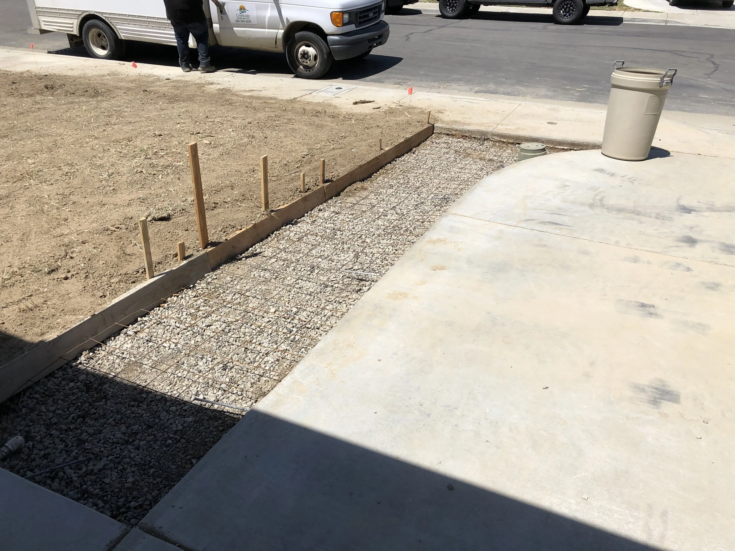 Finished concrete sidewalk with a section of rebar and gravel, next to dirt ground, on a sunny day near parked vans and a trash can.