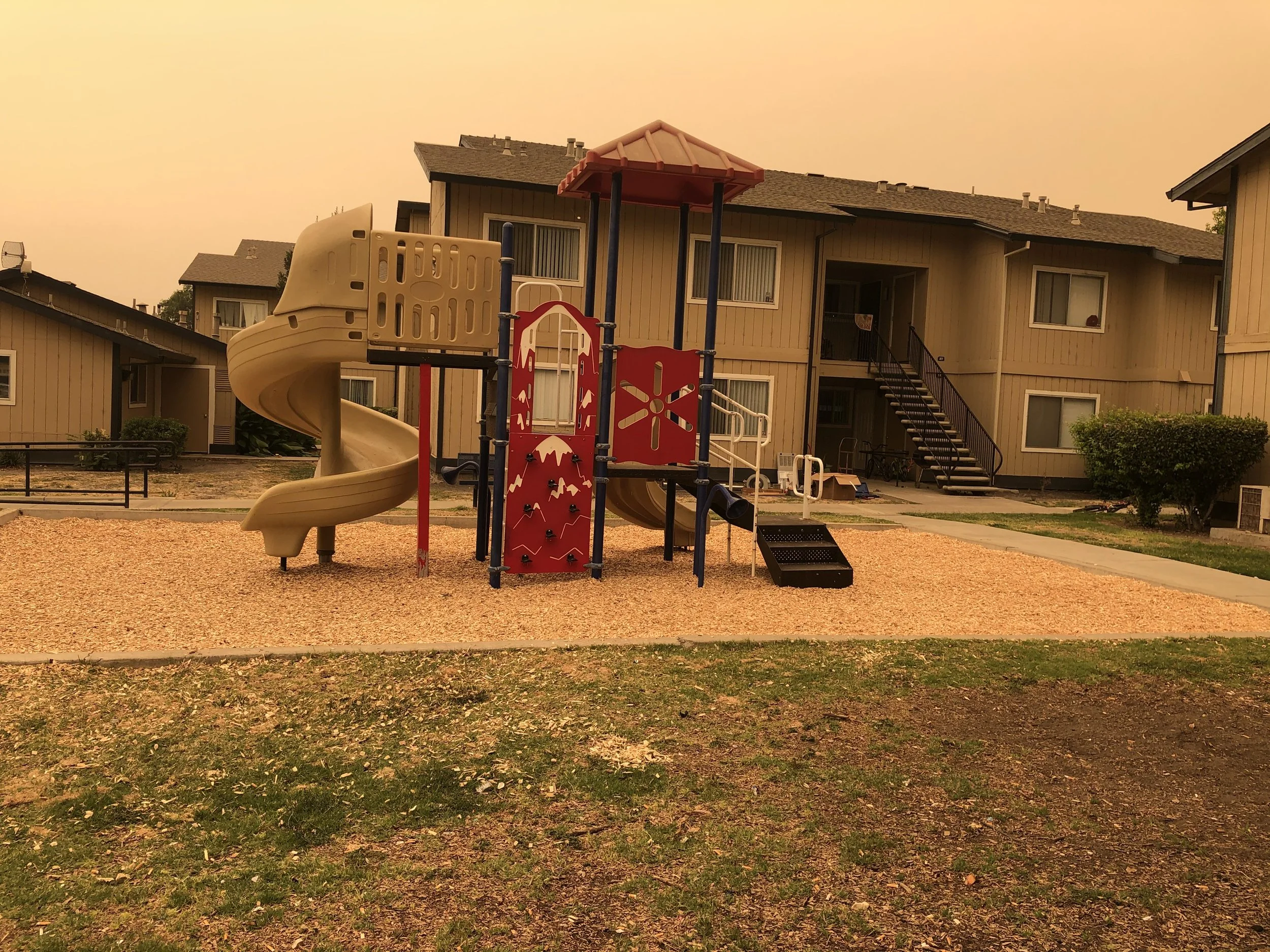 Children's playground with a slide, climbing structures, and a tan spiral slide, located in a residential community with apartment buildings in the background, under an orange sky.