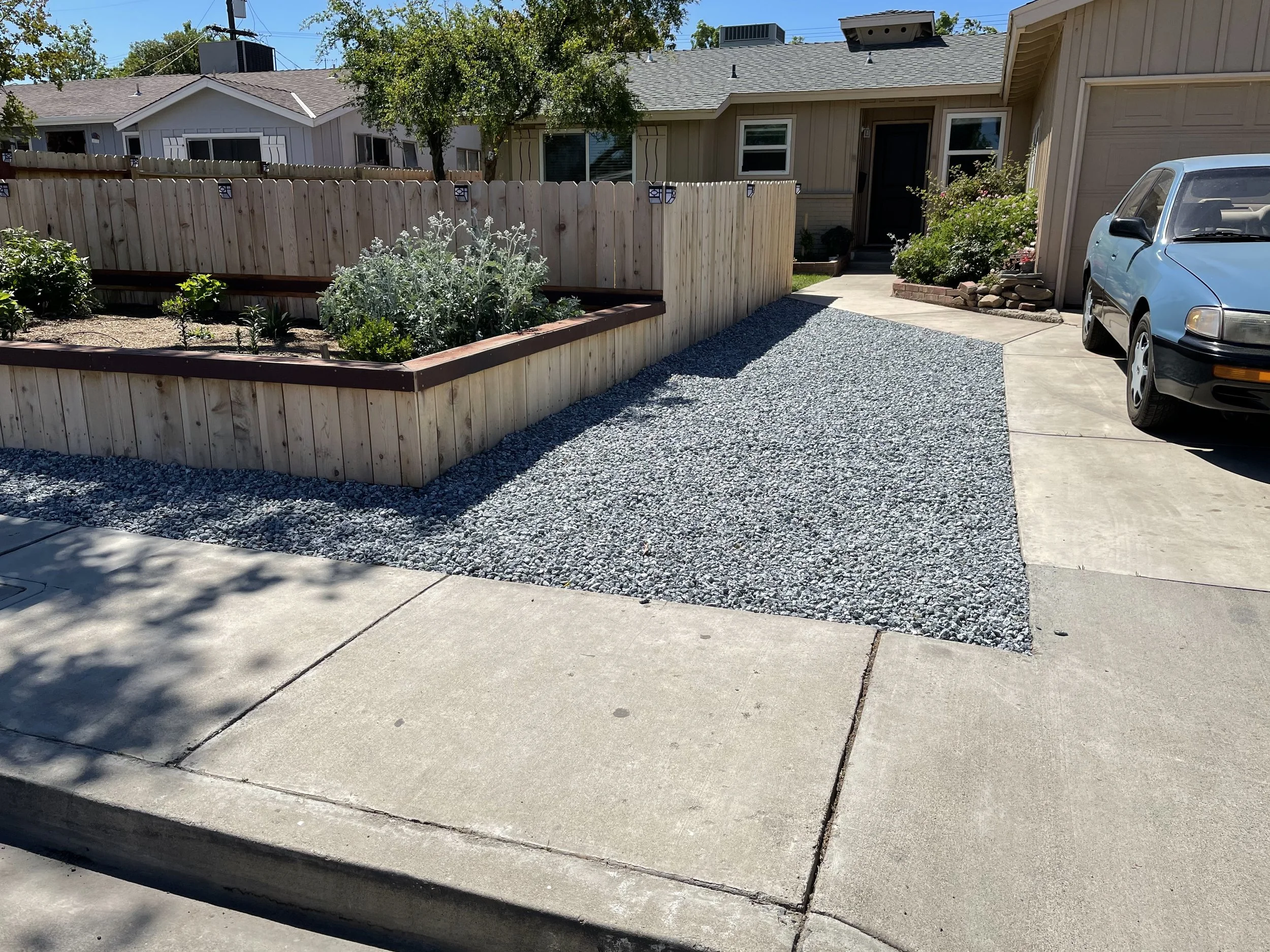 A gravel driveway leading to a house with a parked blue car. There is a wooden fence and a raised garden bed with plants and flowers on the left side. The house has beige siding and a garage door on the right.