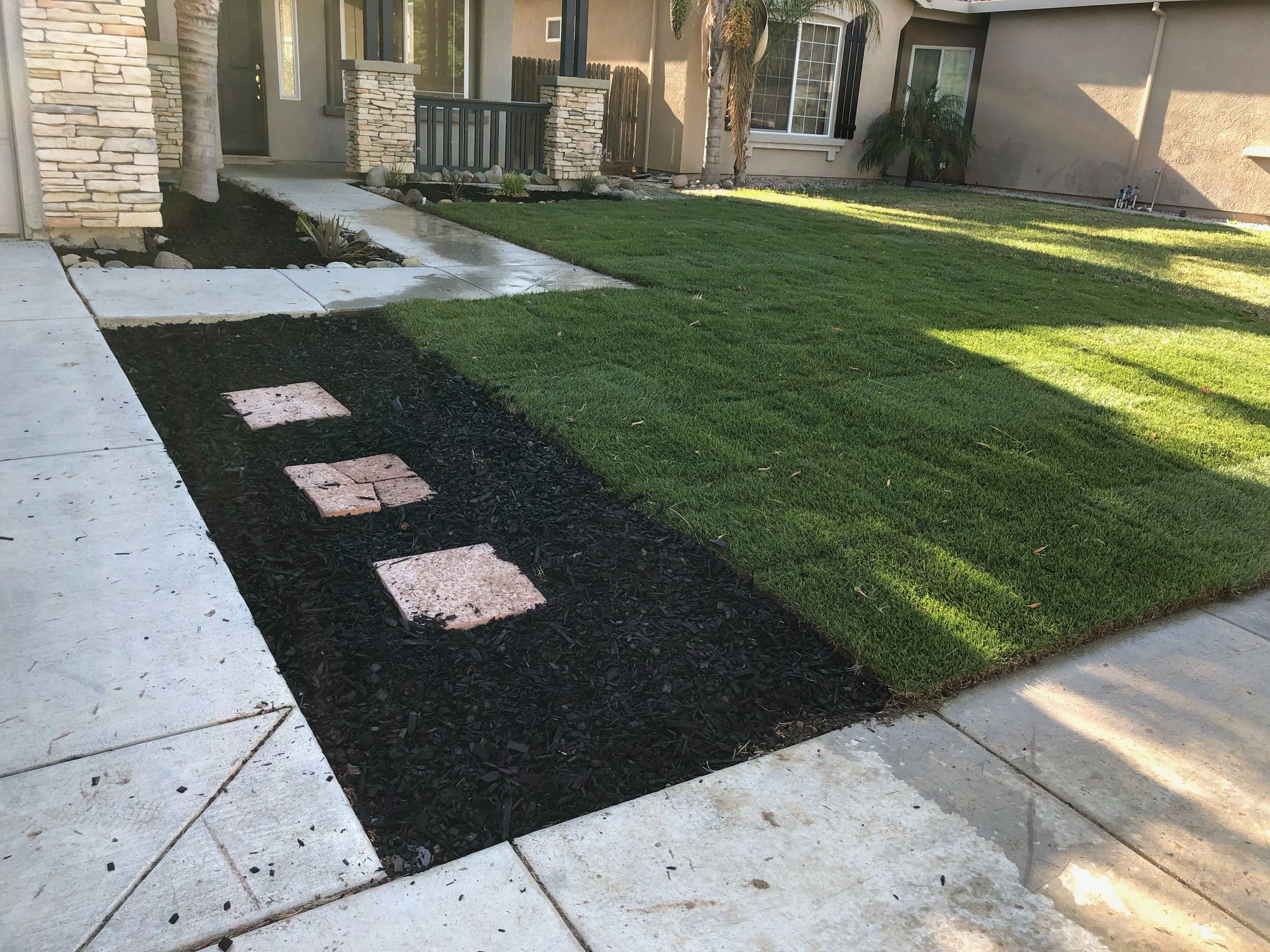A suburban front yard with a concrete walkway, a patch of green grass, and a small garden area with mulch and plants. The house has stone accents, a porch, and palm trees.