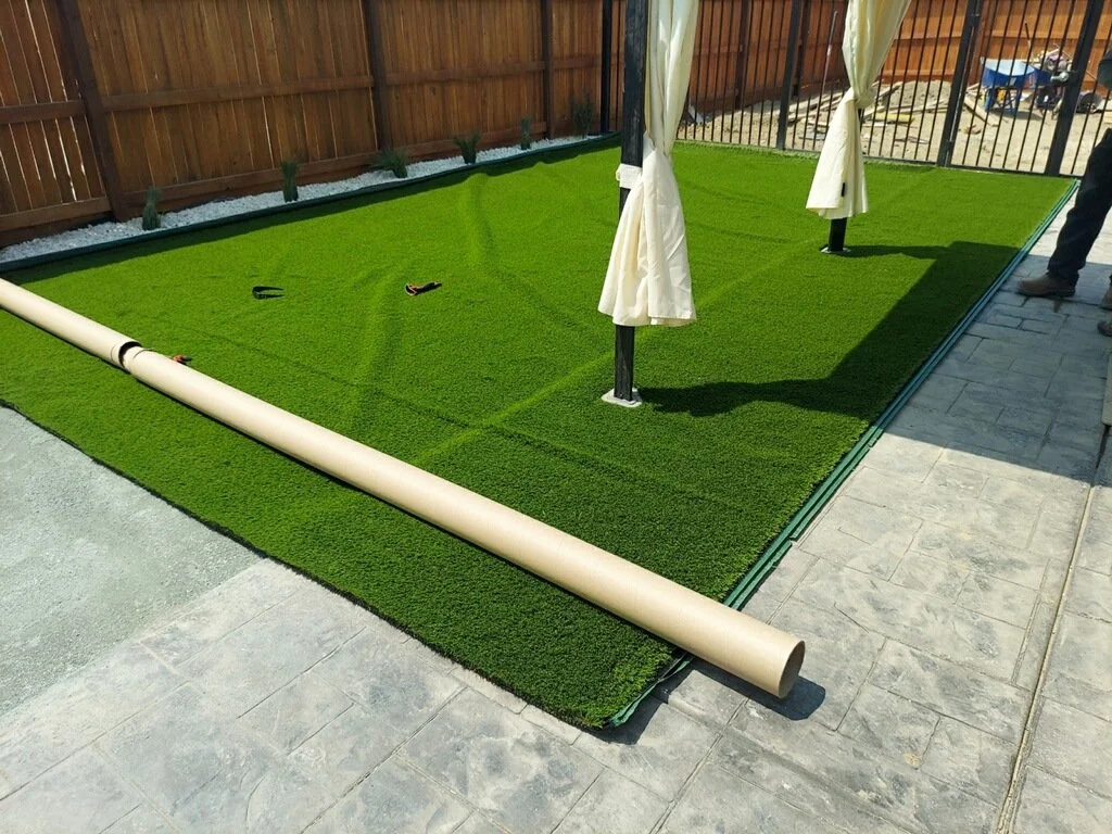 Artificial turf being installed in a backyard with a wooden fence and patio area, with some tools and white fabric curtains standing nearby.