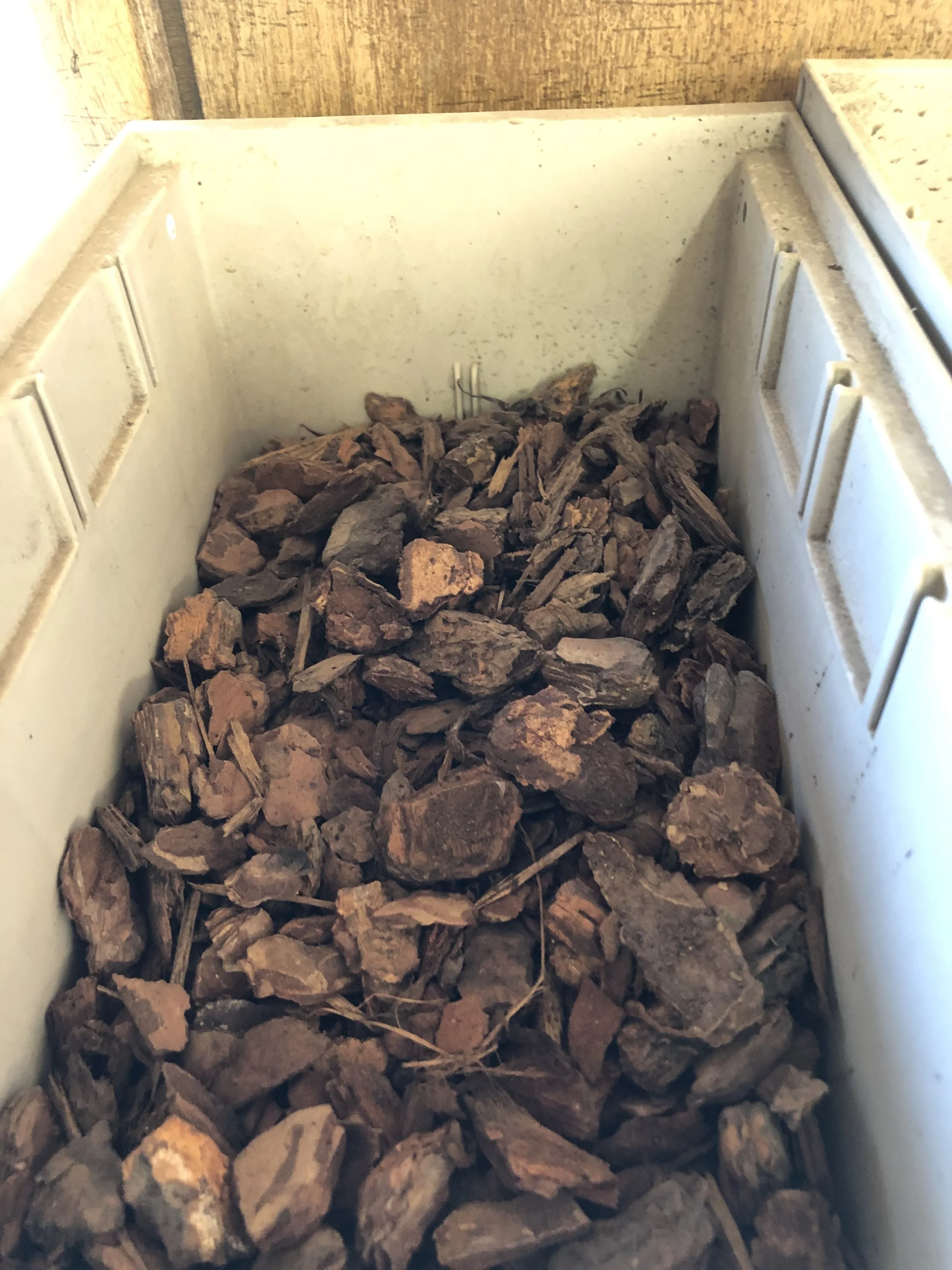 A white bin filled with dark brown rocks or bark chips, with a wooden wall in the background.