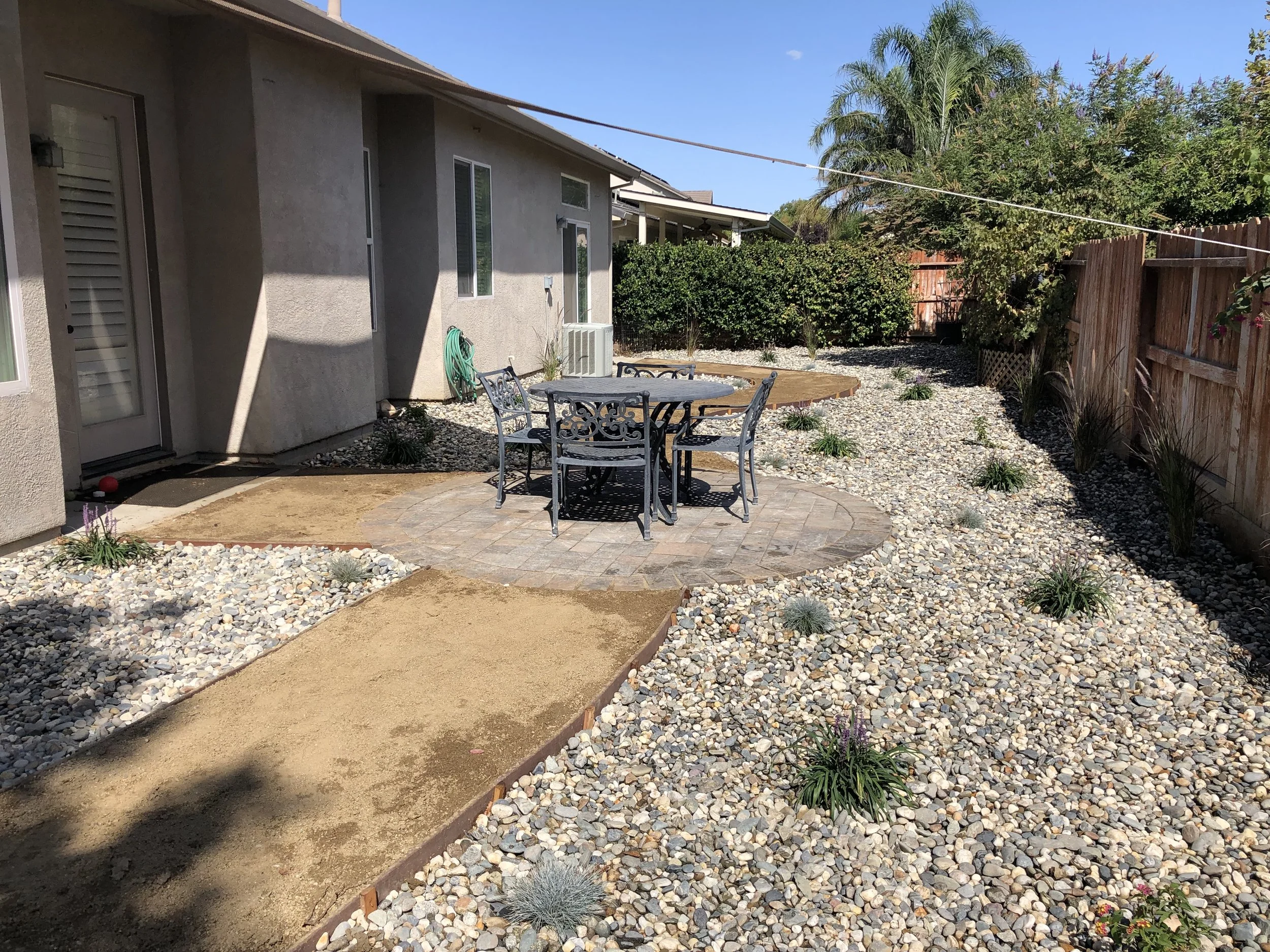 Backyard with a patio area featuring a round table and six chairs, surrounded by gravel and small plants, with a wooden fence and trees in the background.
