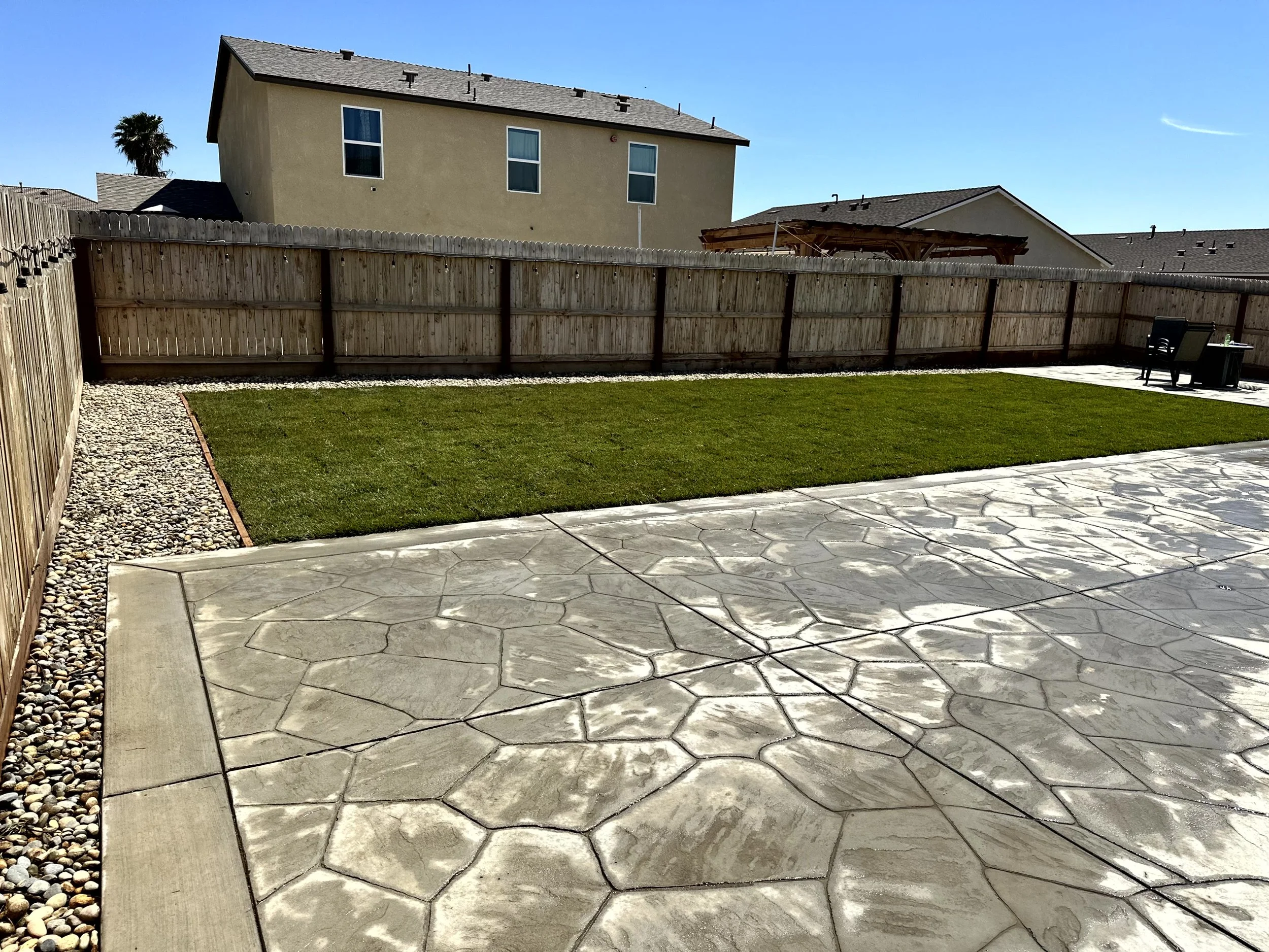 A backyard with a concrete patterned patio, a green lawn, wooden fencing, a small gravel border, and outdoor seating in the corner under a small canopy, with neighboring houses and a clear blue sky in the background.