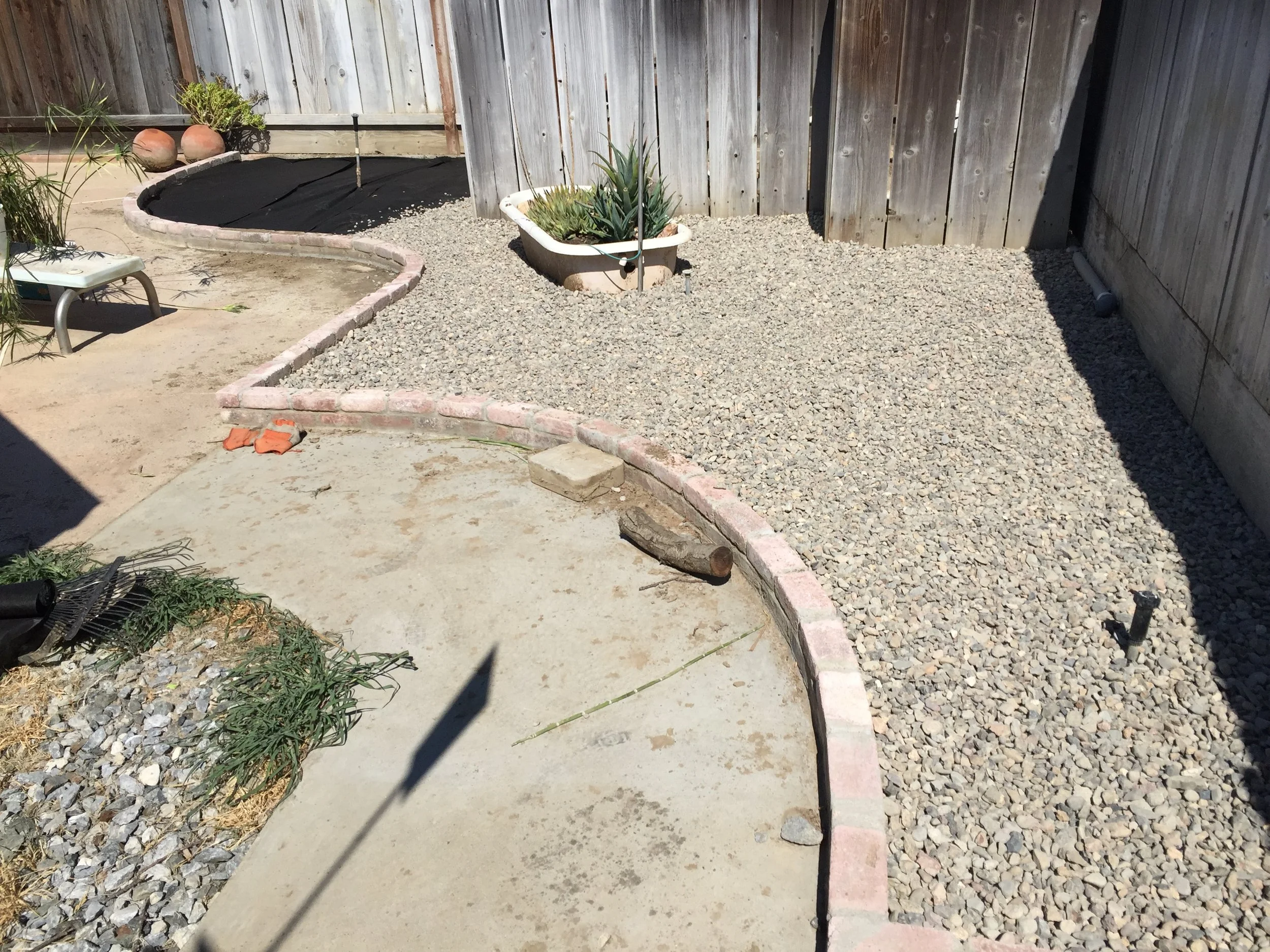 Partially completed backyard landscaping with a curved brick border, gravel, a potted plant, and construction materials.