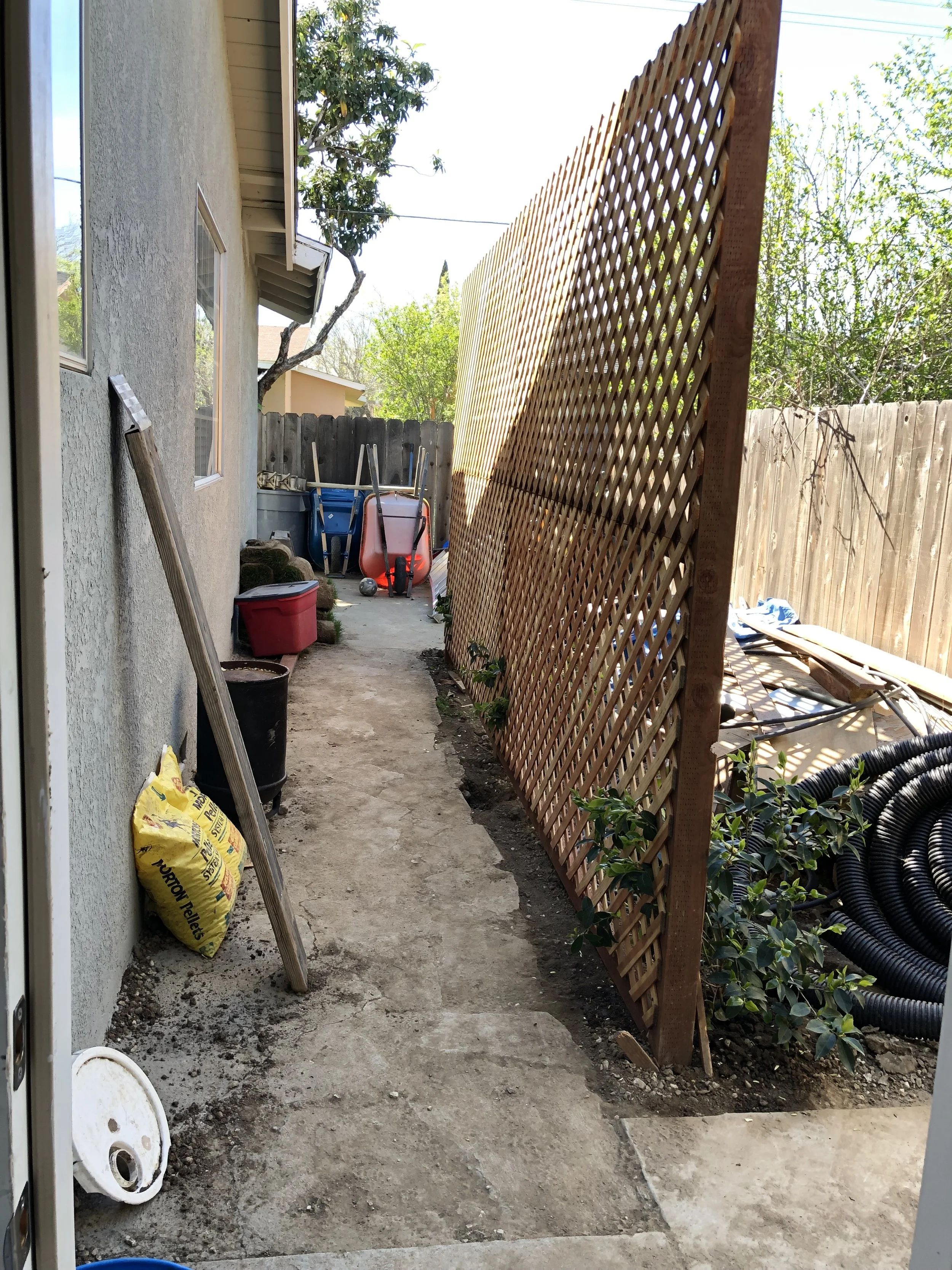 A narrow outdoor backyard space with a dirt pathway, side of a house on the left, and a lattice fence on the right. There are gardening supplies, a red bin, a black barrel, and stacked outdoor furniture in the background. The area is shaded and sunny