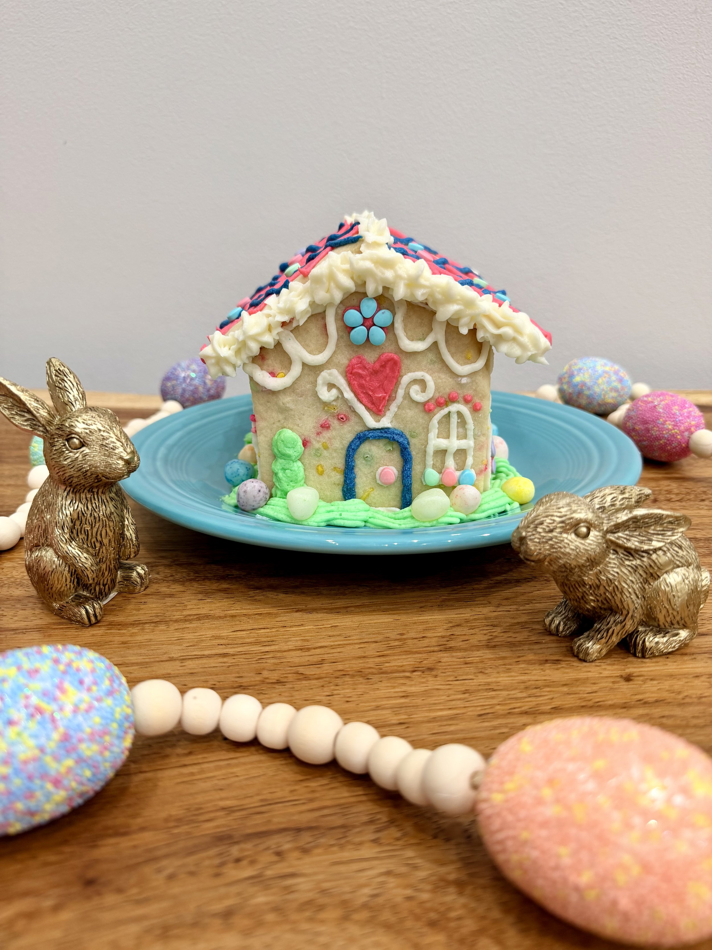 Easter sugar cookie house decorated with piped buttercream grass, jelly bean eggs, pastel icing details, and bunny accents on a blue plate.