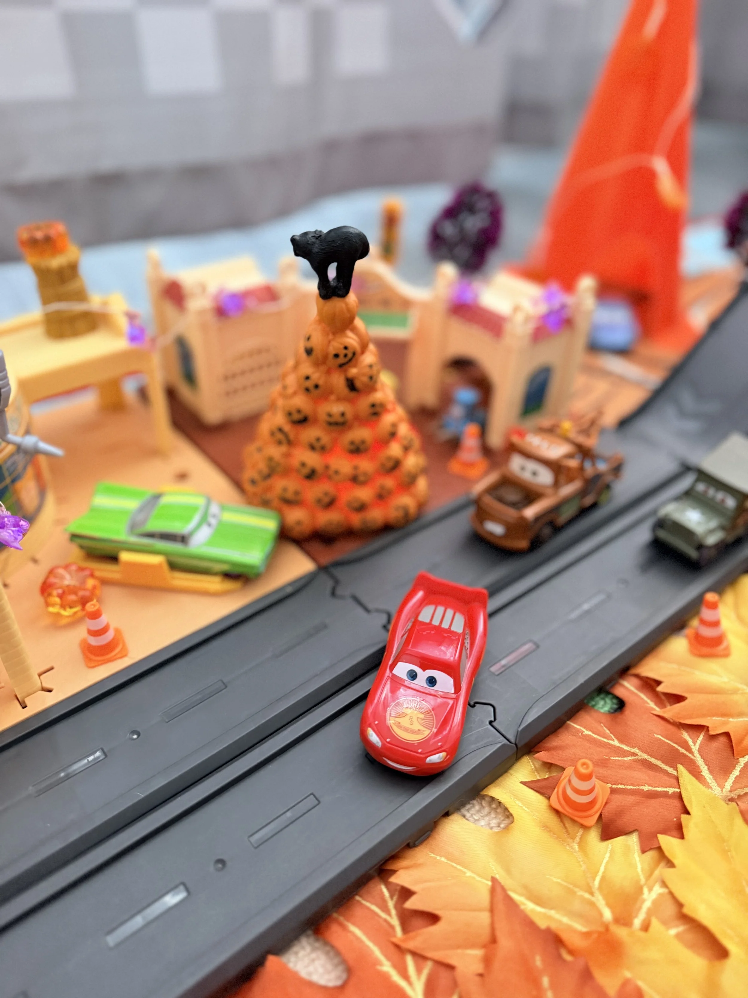 Lightning McQueen drives through a Halloween-decorated Radiator Springs playset with a pumpkin tree, Mater, fall leaves and orange traffic cones.