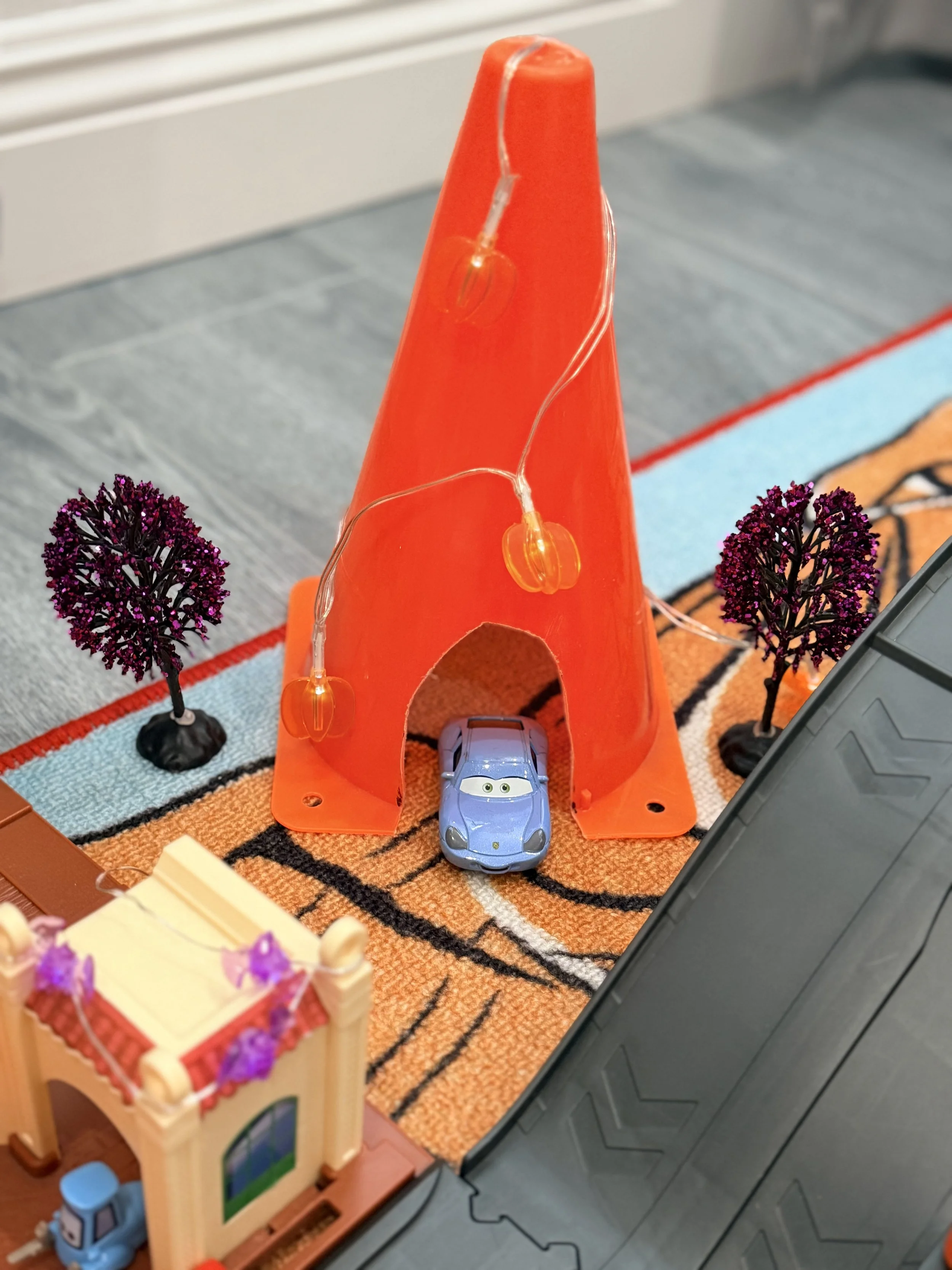 Orange traffic cone turned into a Halloween decoration with pumpkin string lights and miniature purple trees beside a Cars playset.