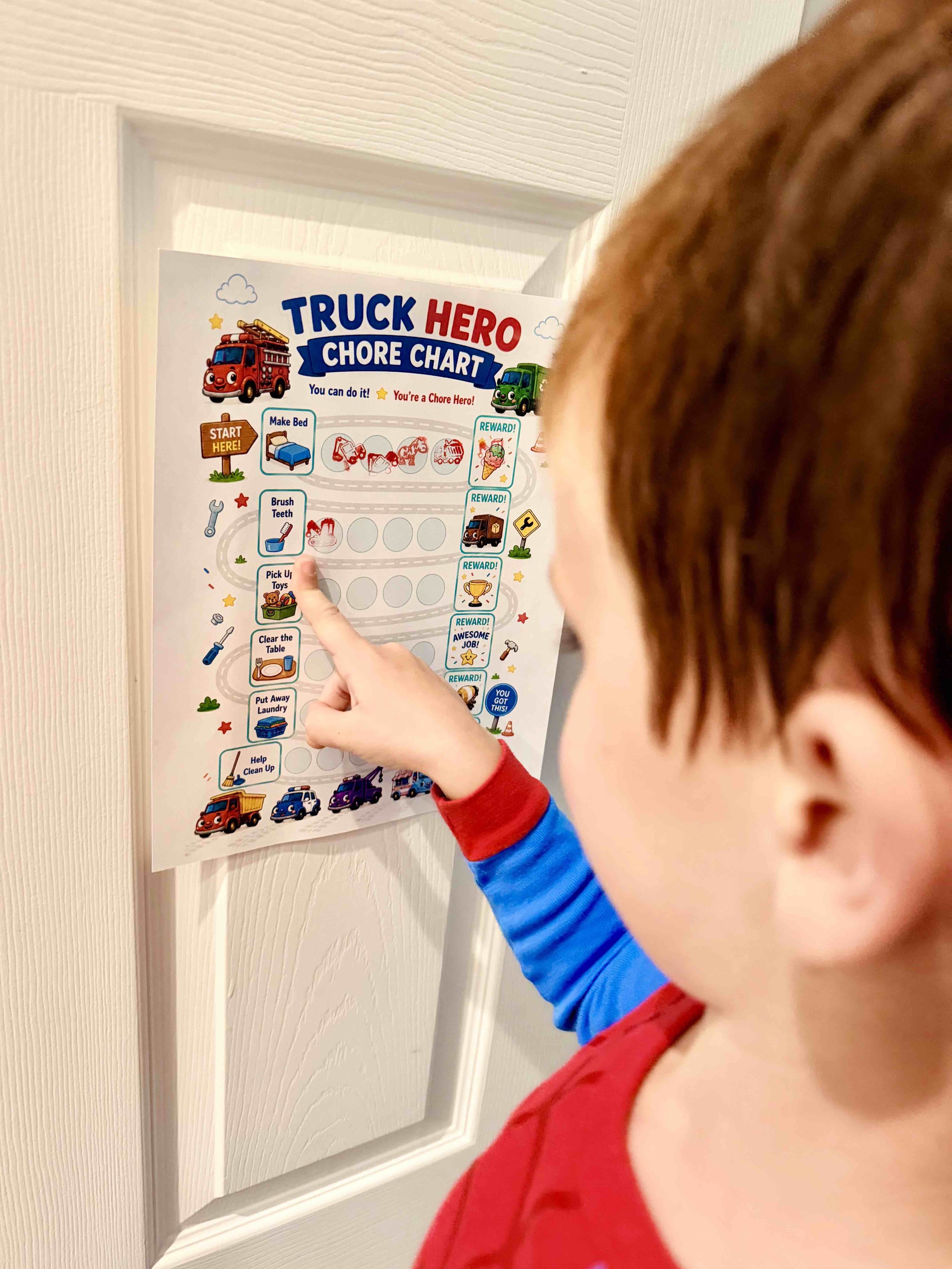 A child pointing at a colorful chore chart on a white door. The chart features small illustrations of trucks and rewards.