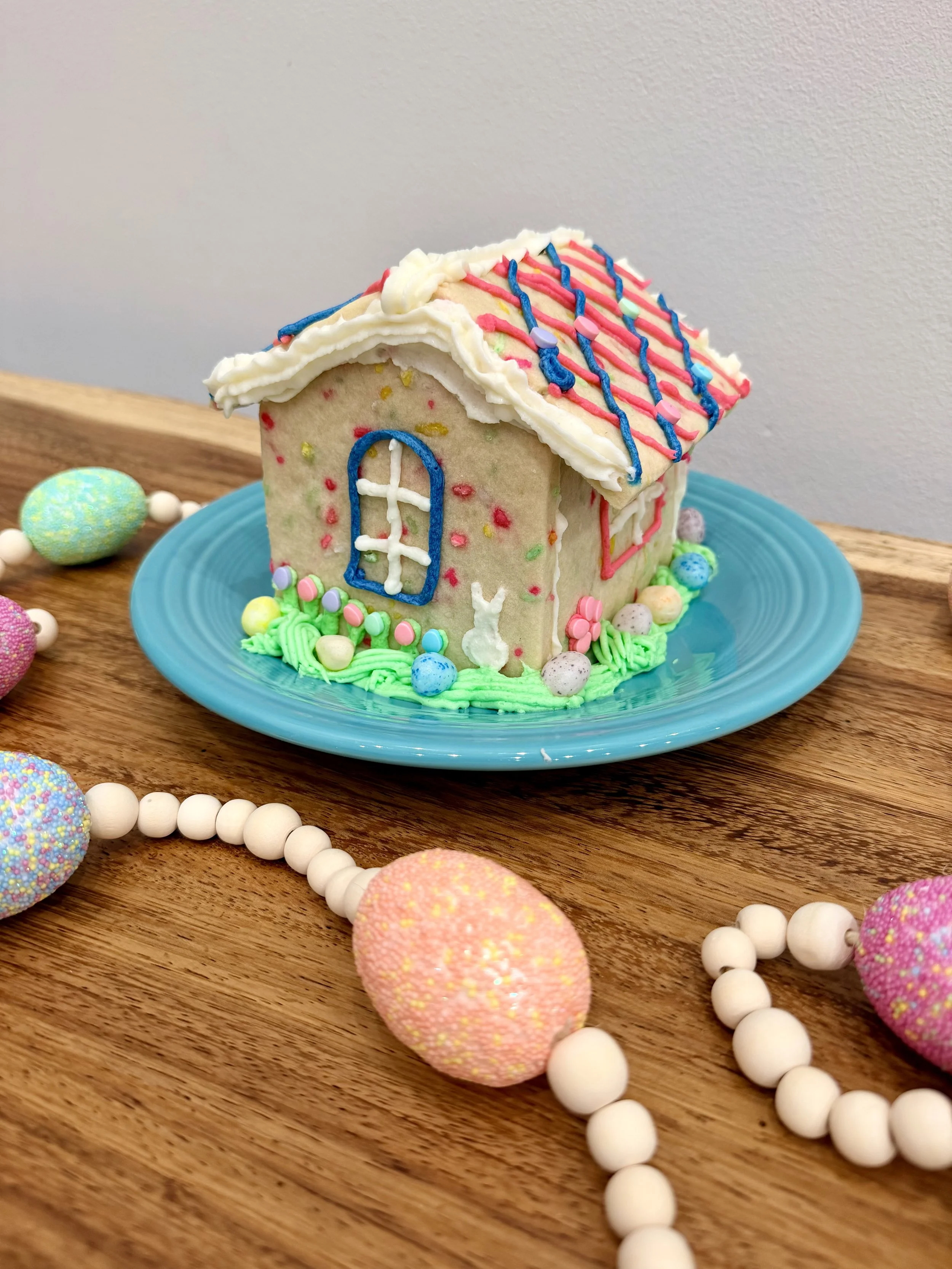 Back of an Easter sugar cookie house with piped buttercream grass, a white bunny silhouette, pastel candy tulips, and jelly bean eggs.