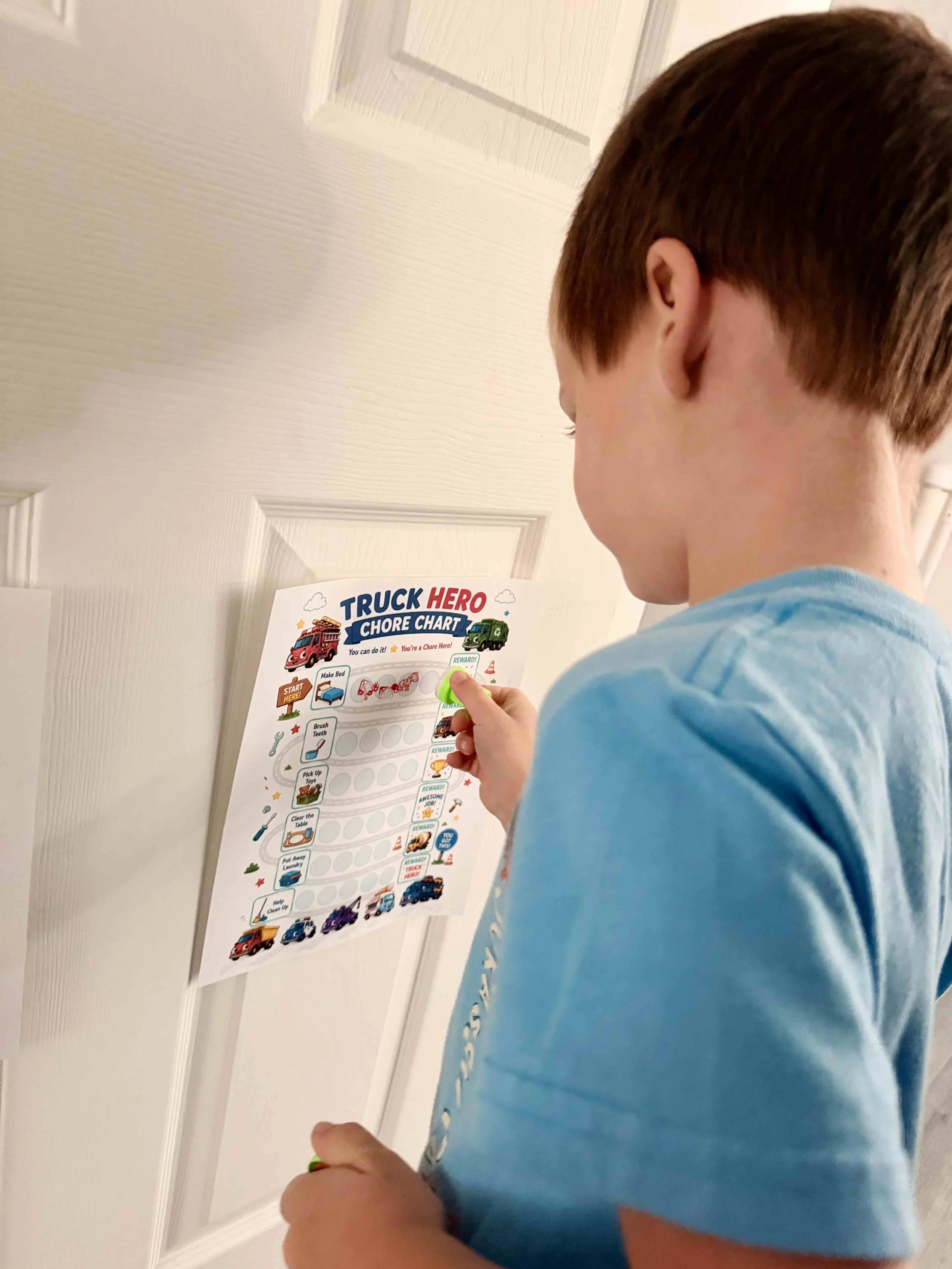 A young boy with reddish hair in a light blue shirt is standing in front of a white door, holding a green marker and working on a colorful chore chart titled 'Truck Hero Chore Chart.'