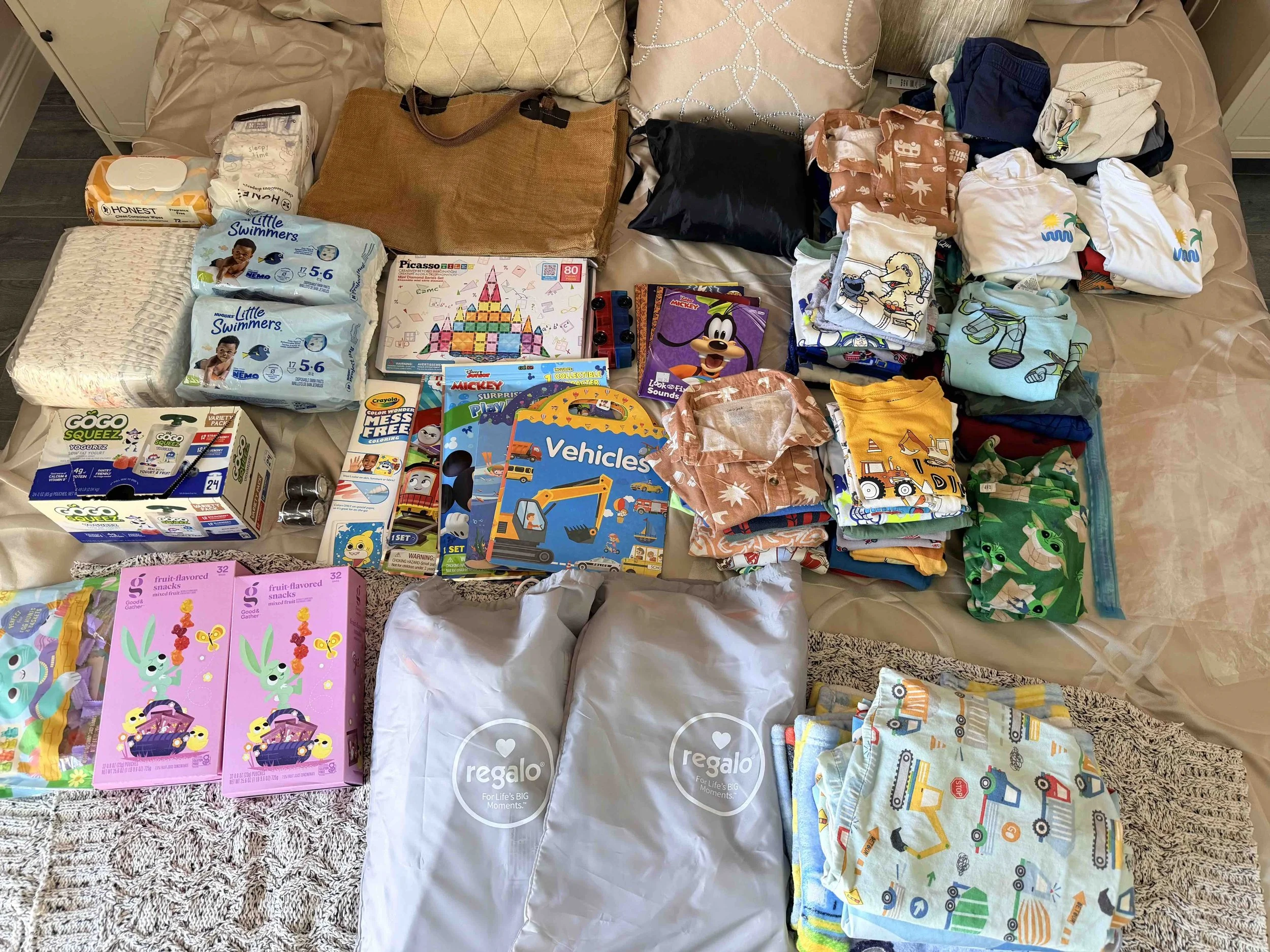 Kids road trip packing checklist with snacks, books, clothes, swim diapers, and toddler travel essentials organized on a bed before a family vacation