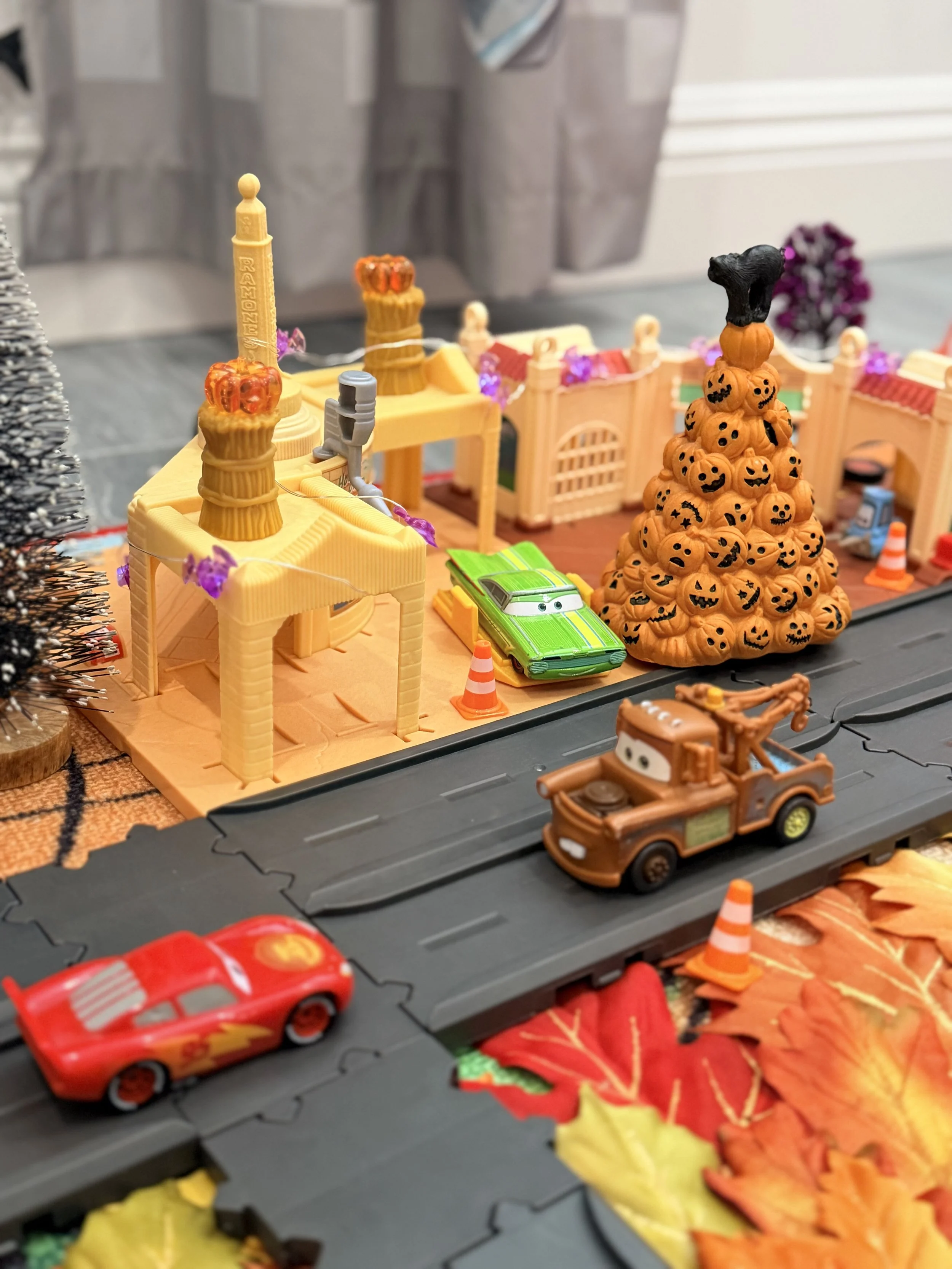 Mater and Lightning McQueen drive past a pumpkin tree in a Halloween Radiator Springs setup with fall leaves, mini pumpkins and purple trees.