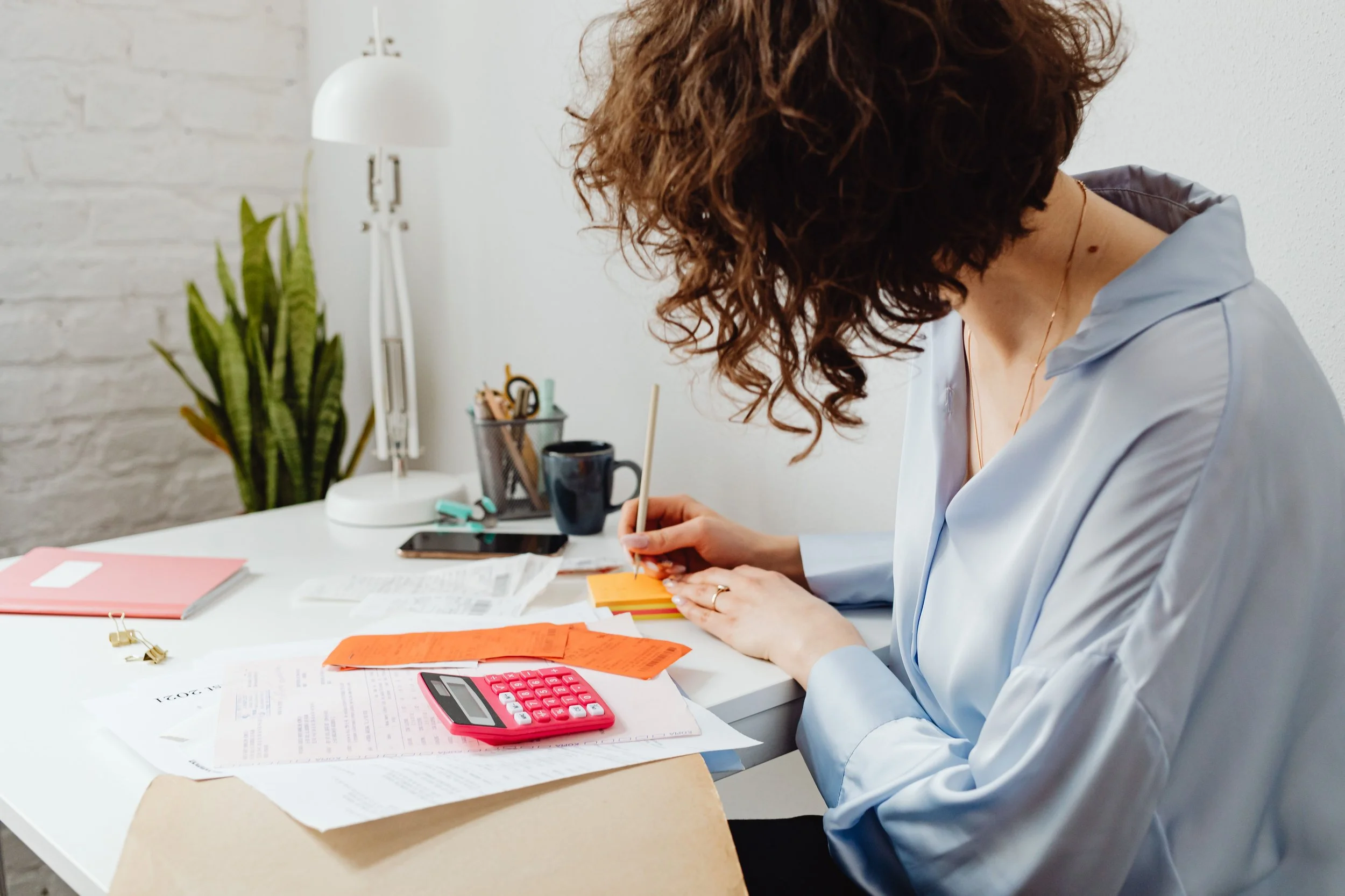 A woman with curly brown hair, wearing a light blue shirt, sitting at a cluttered white desk, writing on orange sticky notes. The desk has various papers, a pink calculator, a black coffee mug, and office supplies. In the background, there's a tall white lamp, a green plant, and a container of stationery items against a white wall.