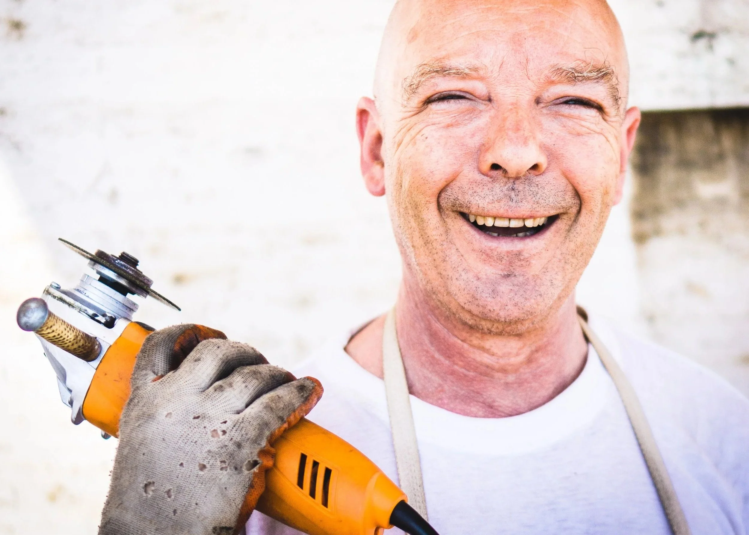 A smiling man holding a torch and a power drill, wearing gloves and a work apron, with a bright background.