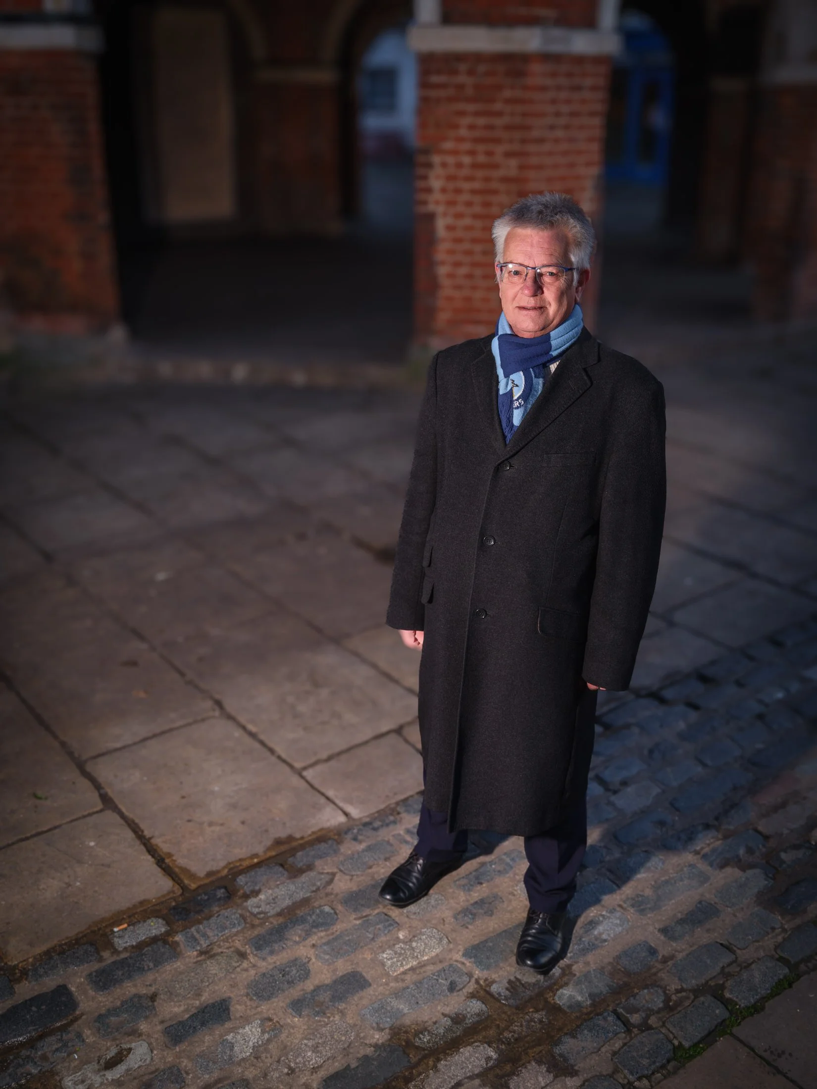 An elderly man standing outdoors in a dark area illuminated by a light. He is wearing glasses, a black coat, a blue scarf, dark pants, and black shoes. He is smiling slightly and looking at the camera. The background features brick structures and a cobblestone street.