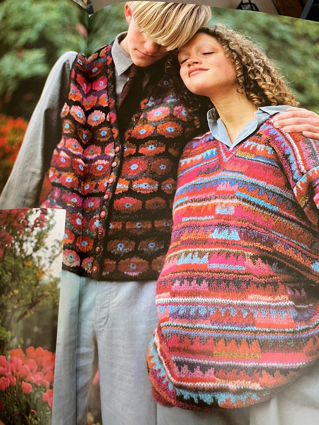 A man and woman are standing close together outdoors, wearing colorful, patterned sweaters. The man has blond hair and is leaning his head on the woman's. The woman has curly hair and is smiling with her eyes closed. There is greenery and flowers in the background.