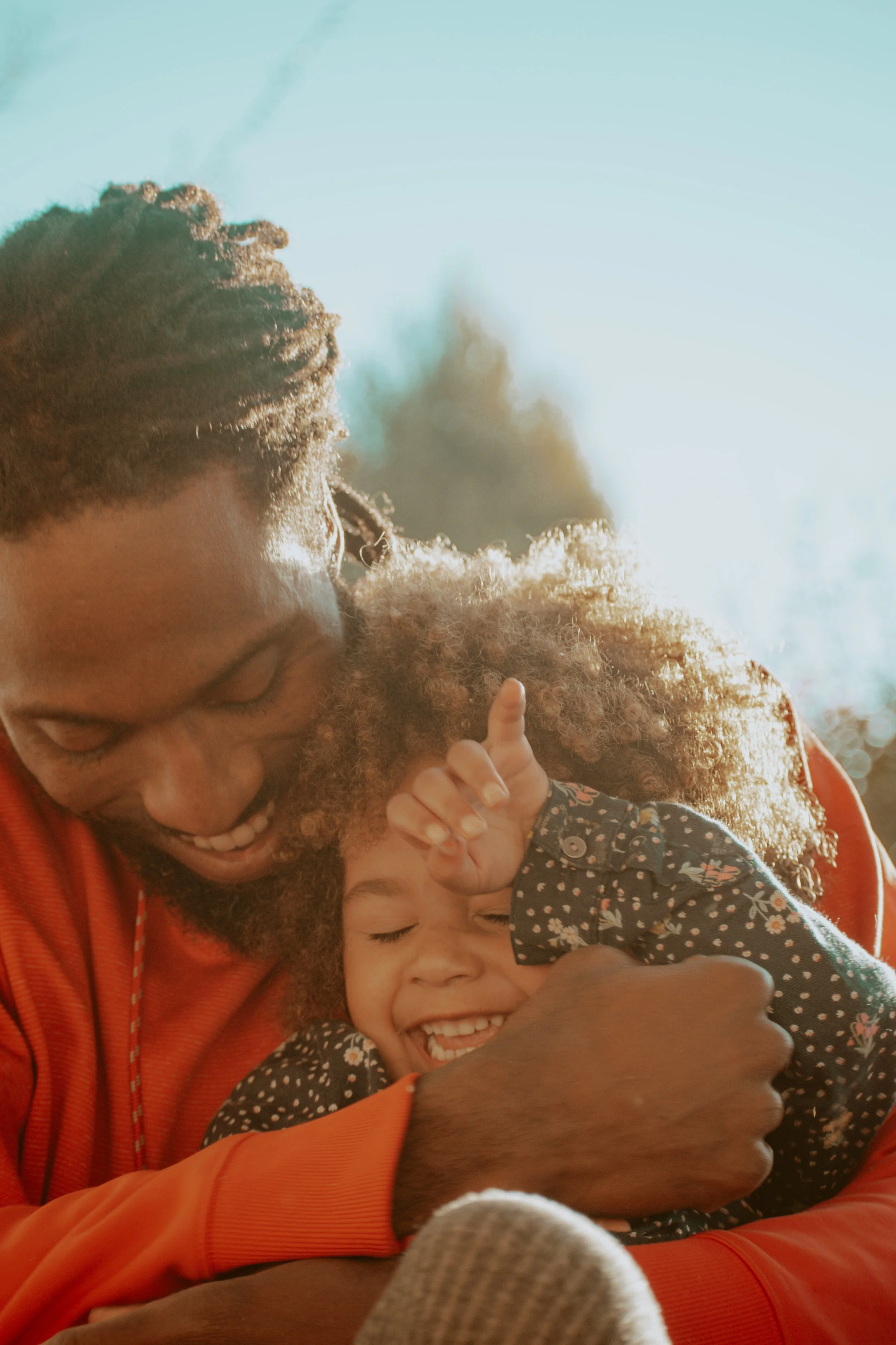A Black father hugs his laughing daughter in golden sunlight, reflecting the hope and healing families find at Neema