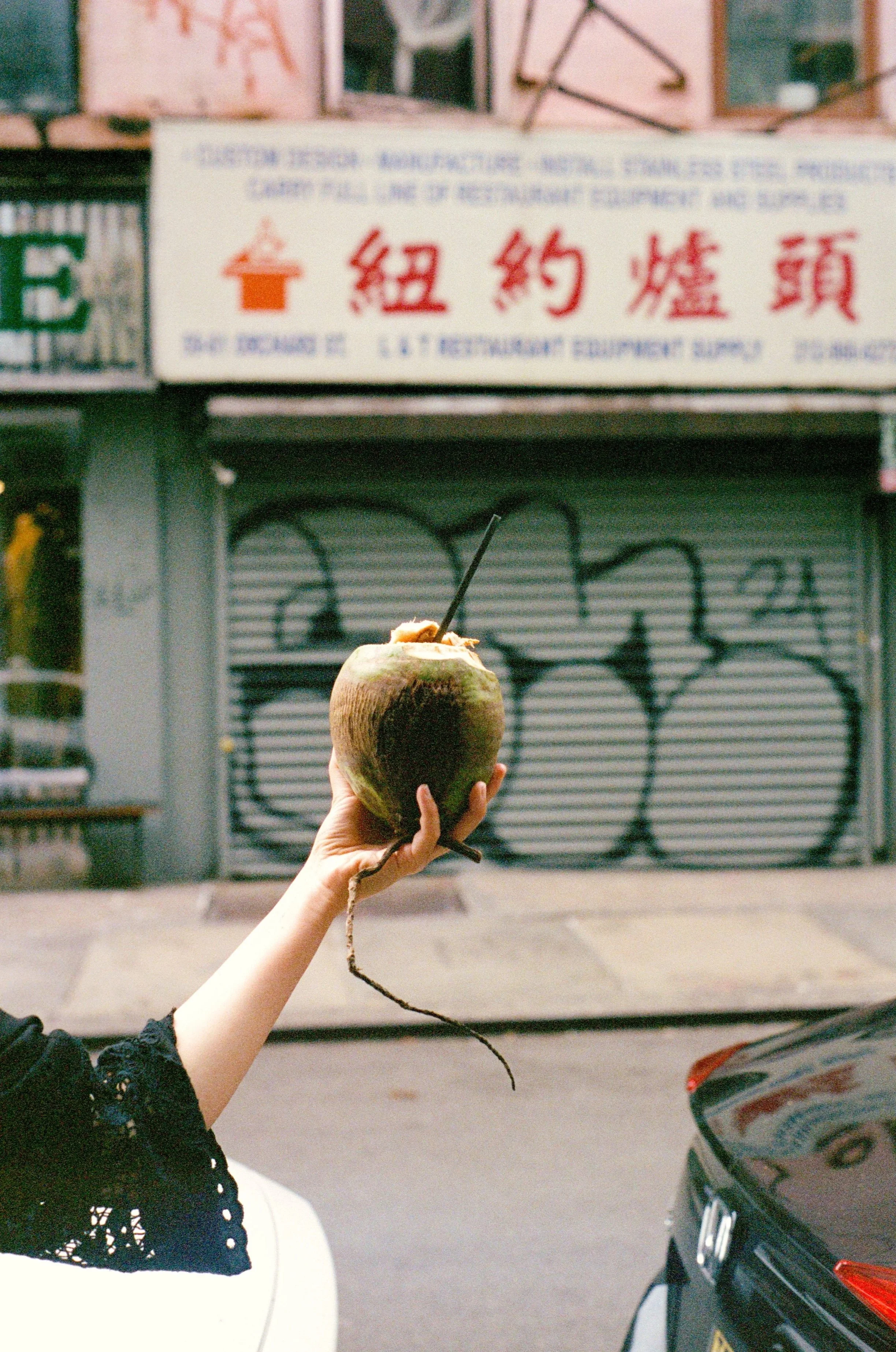 A hand holding a coconut with a straw in it, standing in an urban setting with a closed Chinatown storefront and a car in the background. New York City
