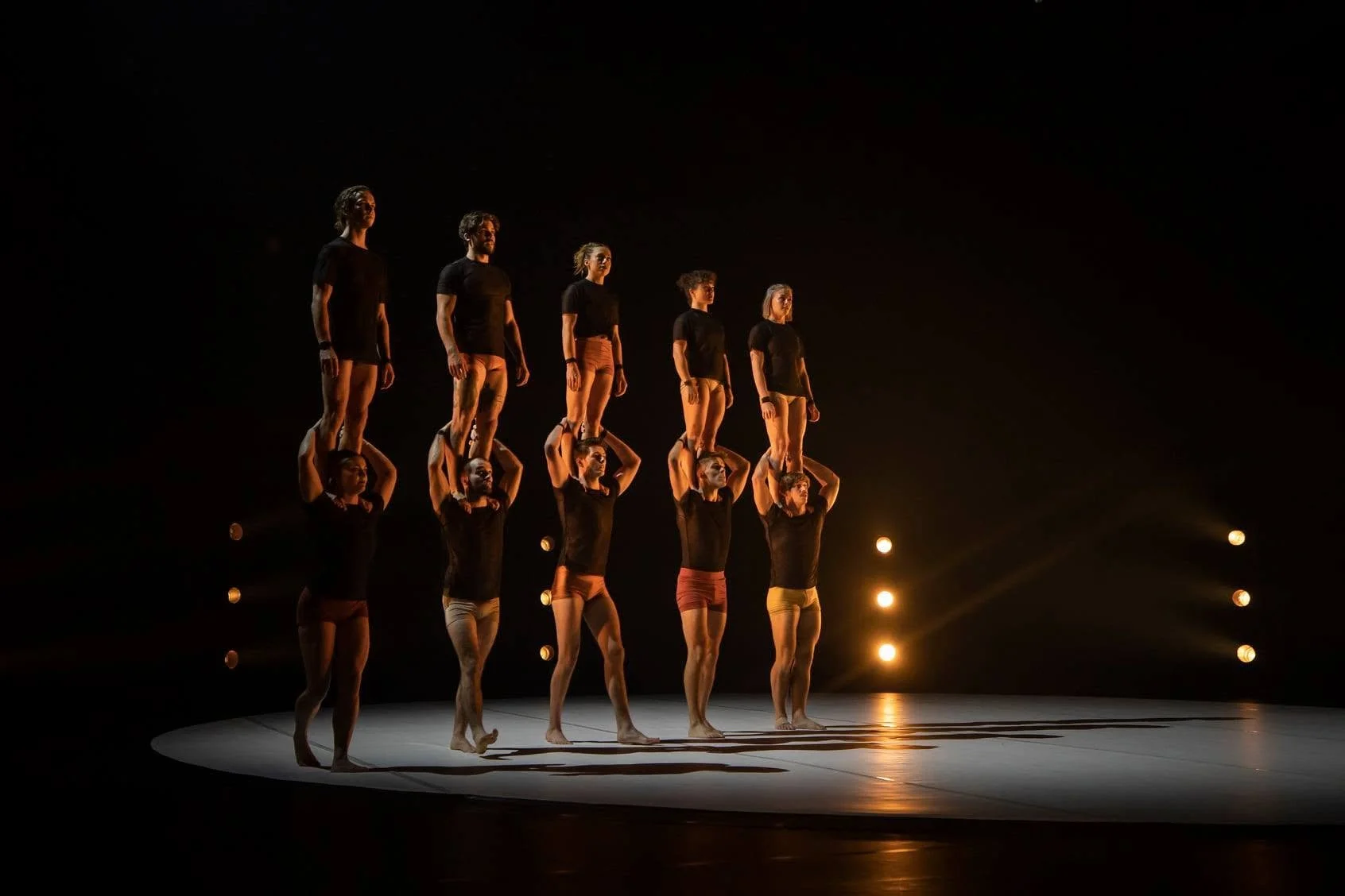 Ten performers in black shirts and colorful shorts balance on top of each other's shoulders in a dark theater, illuminated by stage lights.