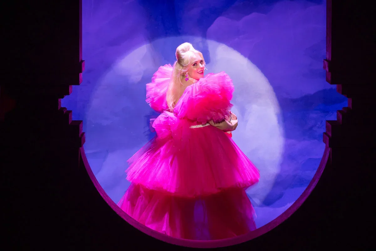 A woman dressed in a bright pink, layered tulle dress with large puffy sleeves, standing on a stage with a blue and purple background, looking over her shoulder.