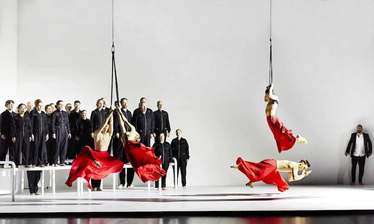 A dance performance with two women on aerial silks and a man on the floor in coordinated red skirts, with backup singers in black standing in the background.