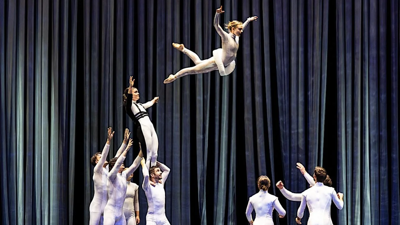 A group of performers in white costumes executing a balancing act on stage, with a curtain background, as one performer is lifted in the air.