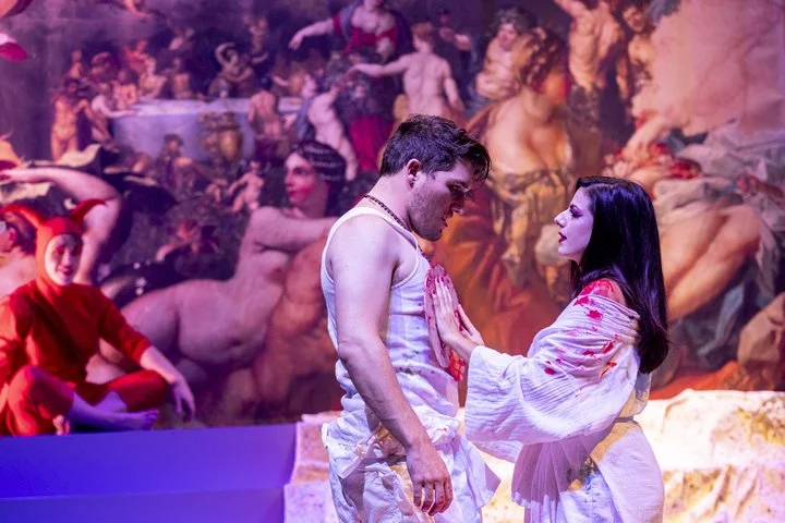 A theatrical performance featuring two actors in the foreground, a male and female, interacting closely. The background is a vibrant, painted scene with multiple other performers in colorful costumes, creating a lively and dynamic atmosphere.