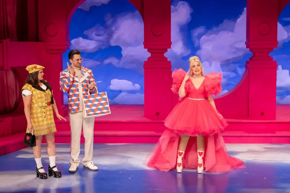 Three actors on stage with a colorful backdrop of clouds and pink archways. The woman on the right wears a bright pink dress with puffed sleeves and heart-patterned socks, speaking animatedly. A man and a woman stand on the left wearing retro-style clothing, listening to her.