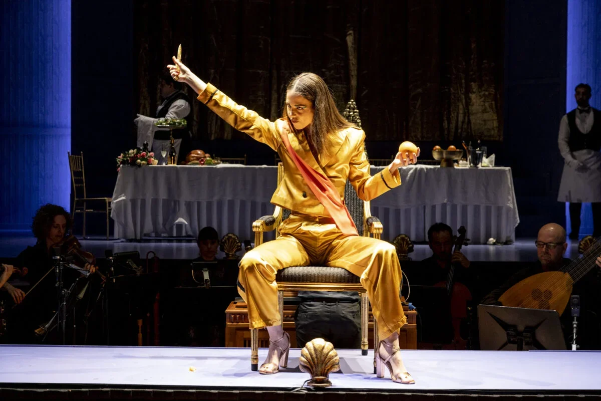 A performer in a shiny gold outfit with a pink sash, sitting on a chair on stage, pointing upwards with a serious expression, holding an apple in one hand. An orchestra is visible behind her, and a table with food and drinks is in the background.