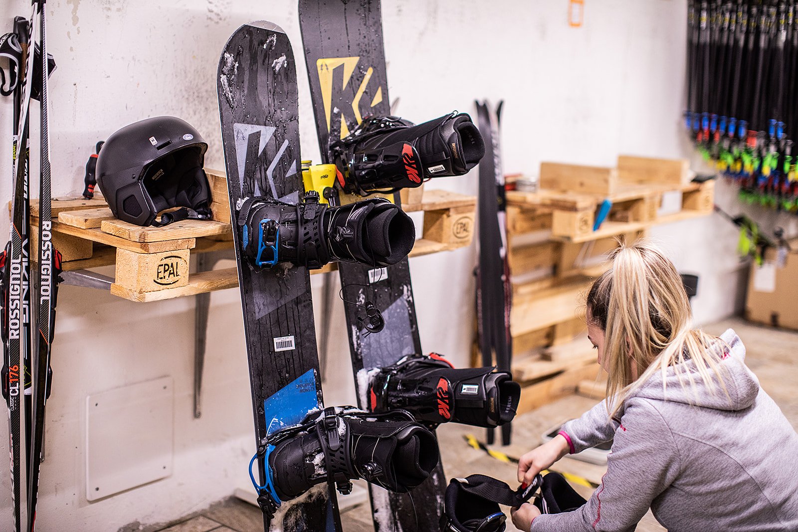 A young woman is organizing ski gear in a ski shop. Ski goggles, helmets, and skis can be seen on a shelf.