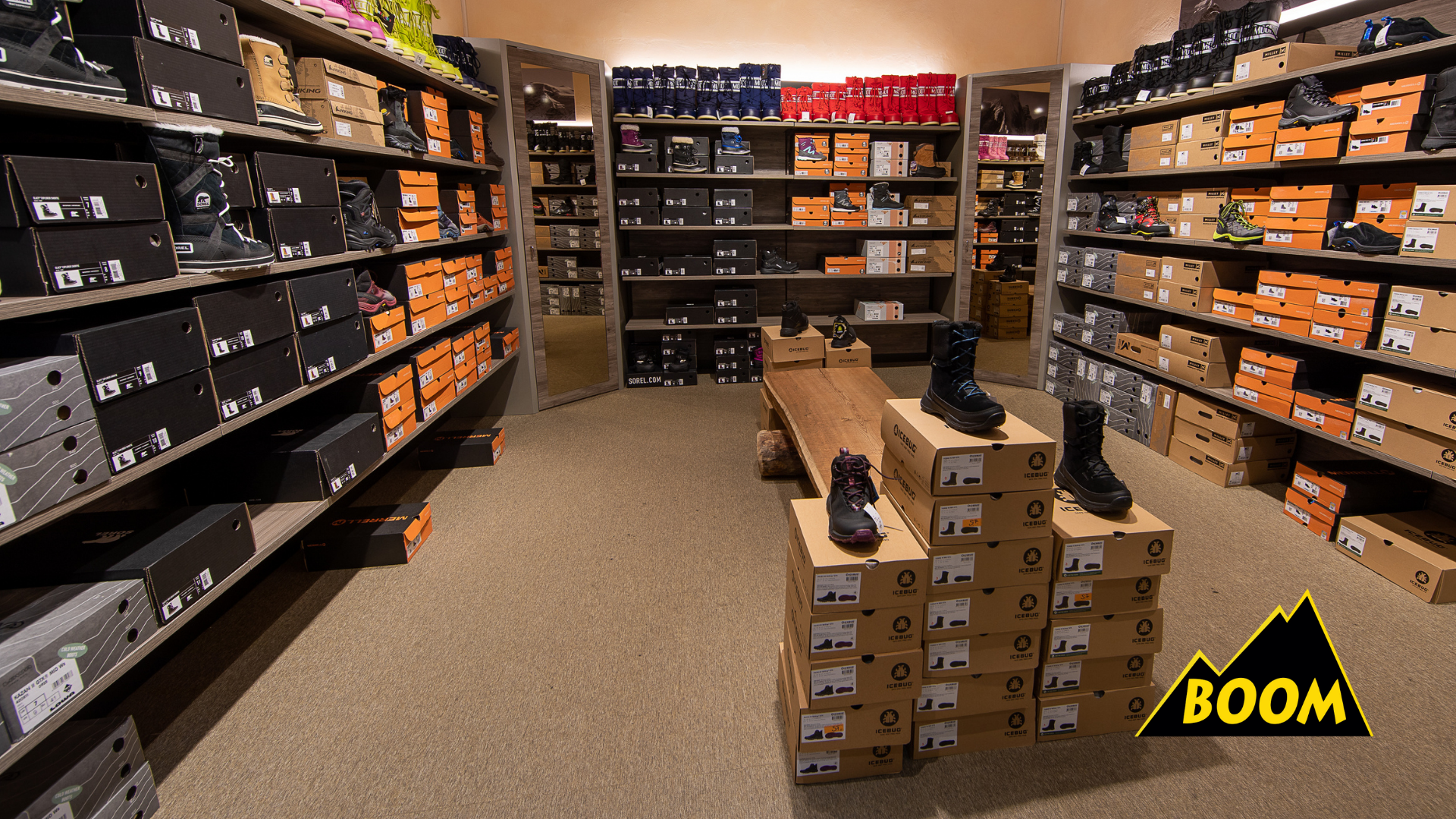 Ladengeschäft für Skischuhe und Wintersportausrüstung mit im Hintergrund gefärbten Schuhen auf Regalen, mittig Schwarz Schuhe auf Kartons und rechts schwarze Schuhe auf Regalen, mit einem Holzpult im Vordergrund.