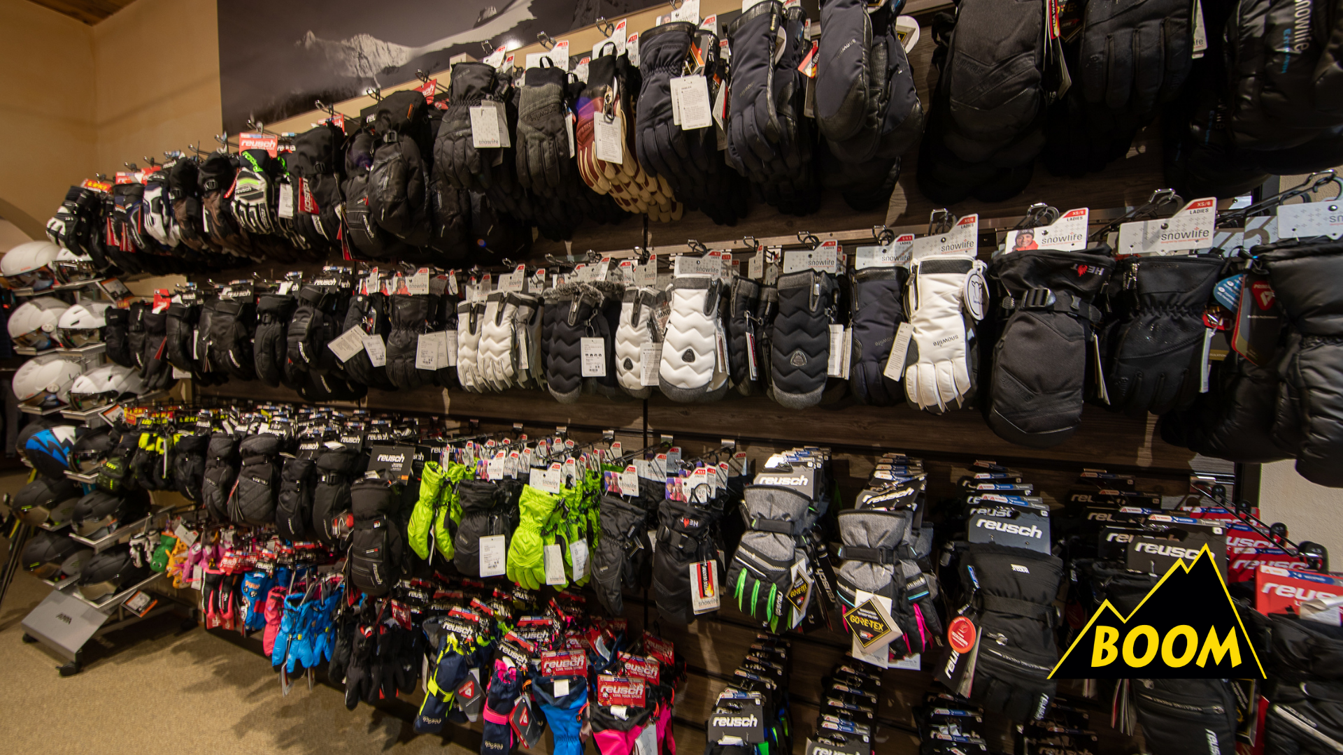 Regale im Sportgeschäft mit Skihandschuhen, Skimaschinen und Helmen. Verschiedene Handschuhe in Schwarz, Weiß, Grau und Neongrün hängen an den Regalen, darunter auch Skibrillen. Es gibt auch Skiausrüstung wie Helme auf der linken Seite.