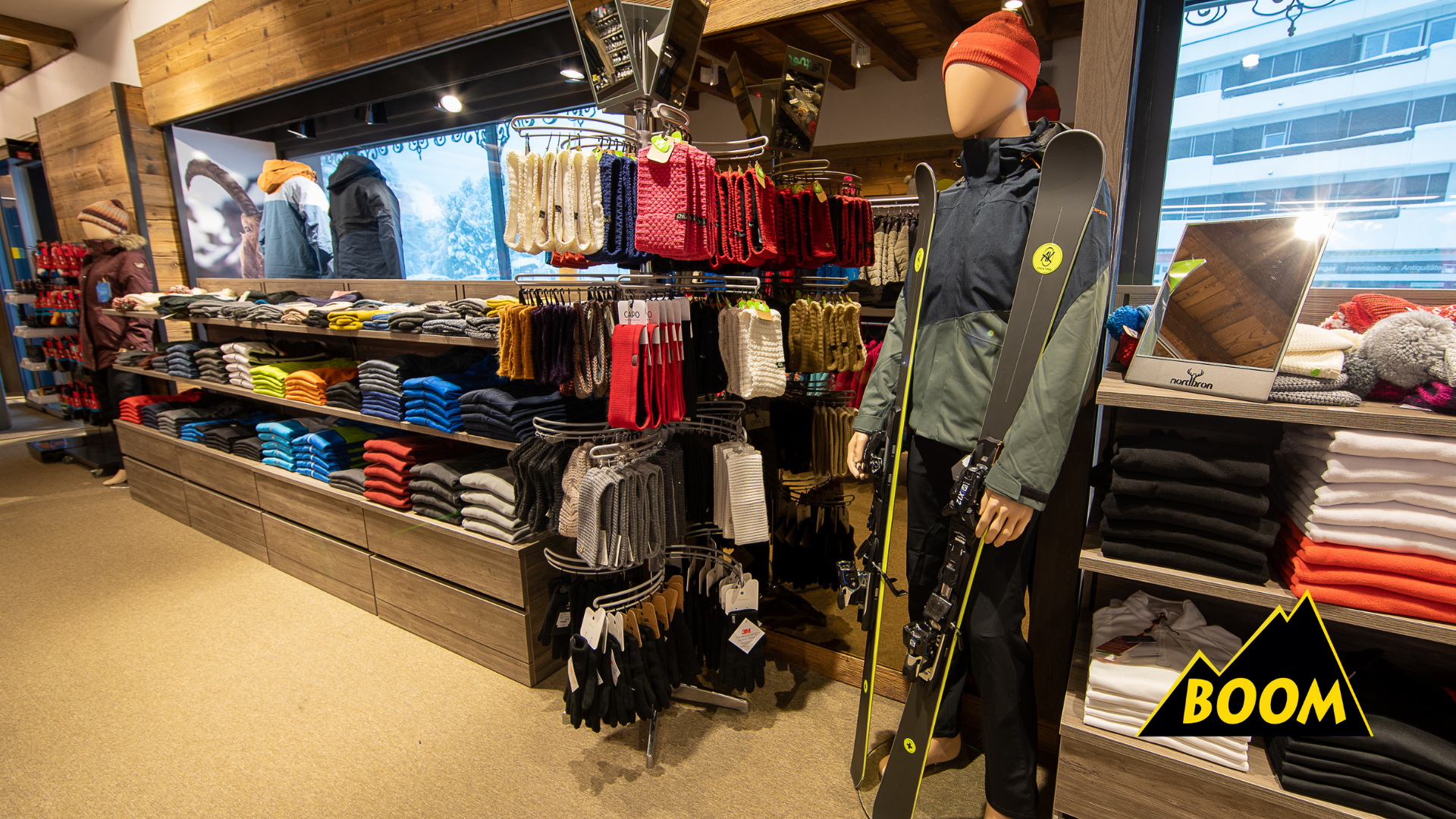 Ein Laden mit Winterkleidung und Skiausrüstung, darunter eine Skijacke und Skier an einem stilisierten Menschen aus Pappmaché. Es sind Regale mit Pullovern, Handschuhen und Mützen, sowie Schneeketten und Skihandschuhe zu sehen.