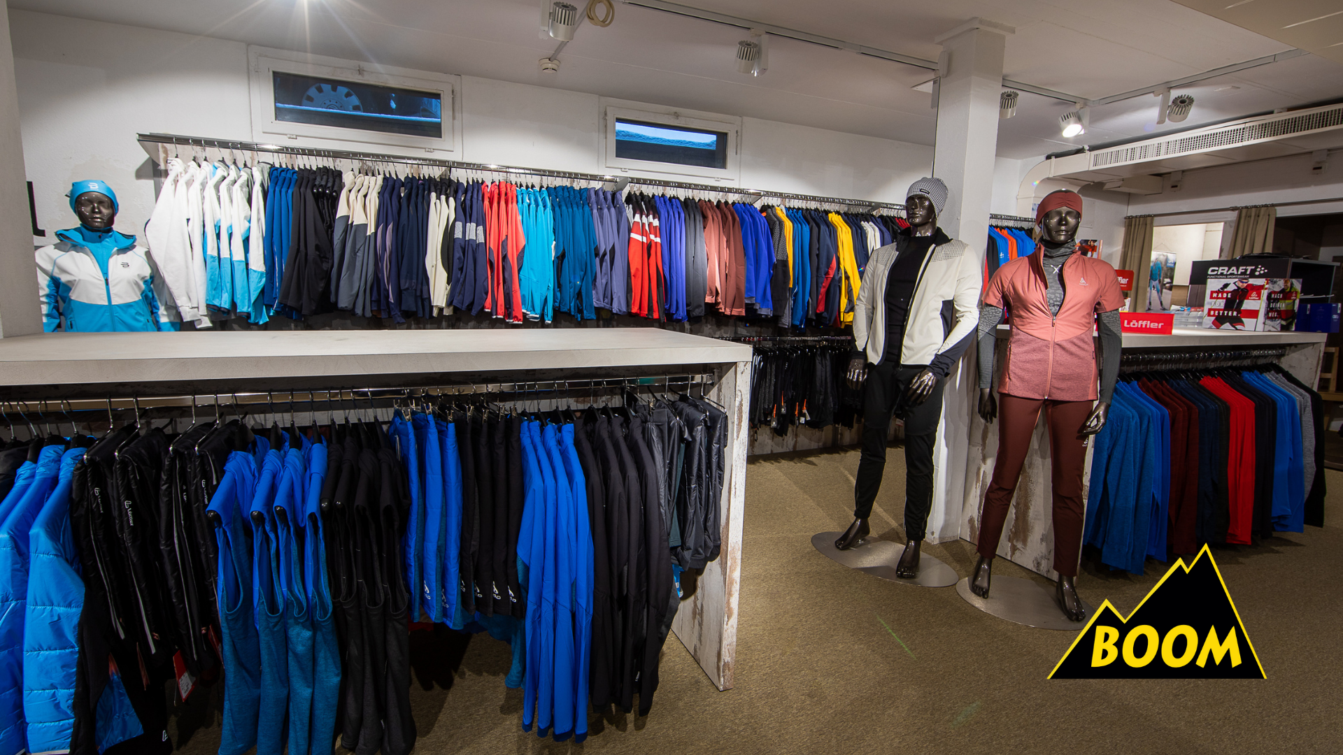 Sportbekleidung in einem Laden, Mannequins in Sportoutfits, Regale mit Jacken und Hosen, Schilder mit Markeninformationen, im Hintergrund eine Kasse, Werbedisplay mit dem Wort 'BOOM' in gelb auf schwarzem Hintergrund.