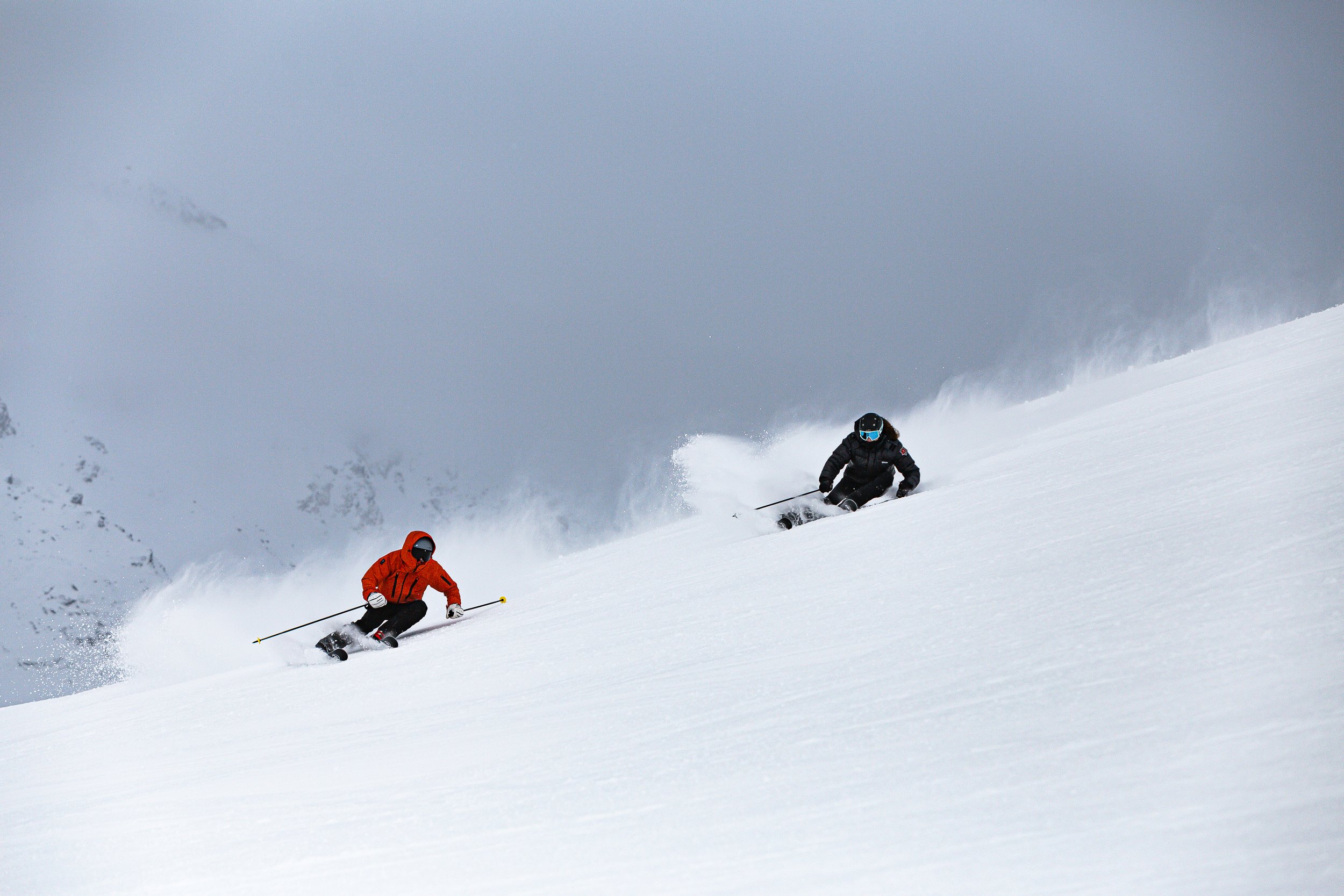 Drei Skifahrer in Winterkleidung und Helm beim Skifahren in tiefem Schnee auf einer Bergpiste, mit bewölktem Himmel im Hintergrund.