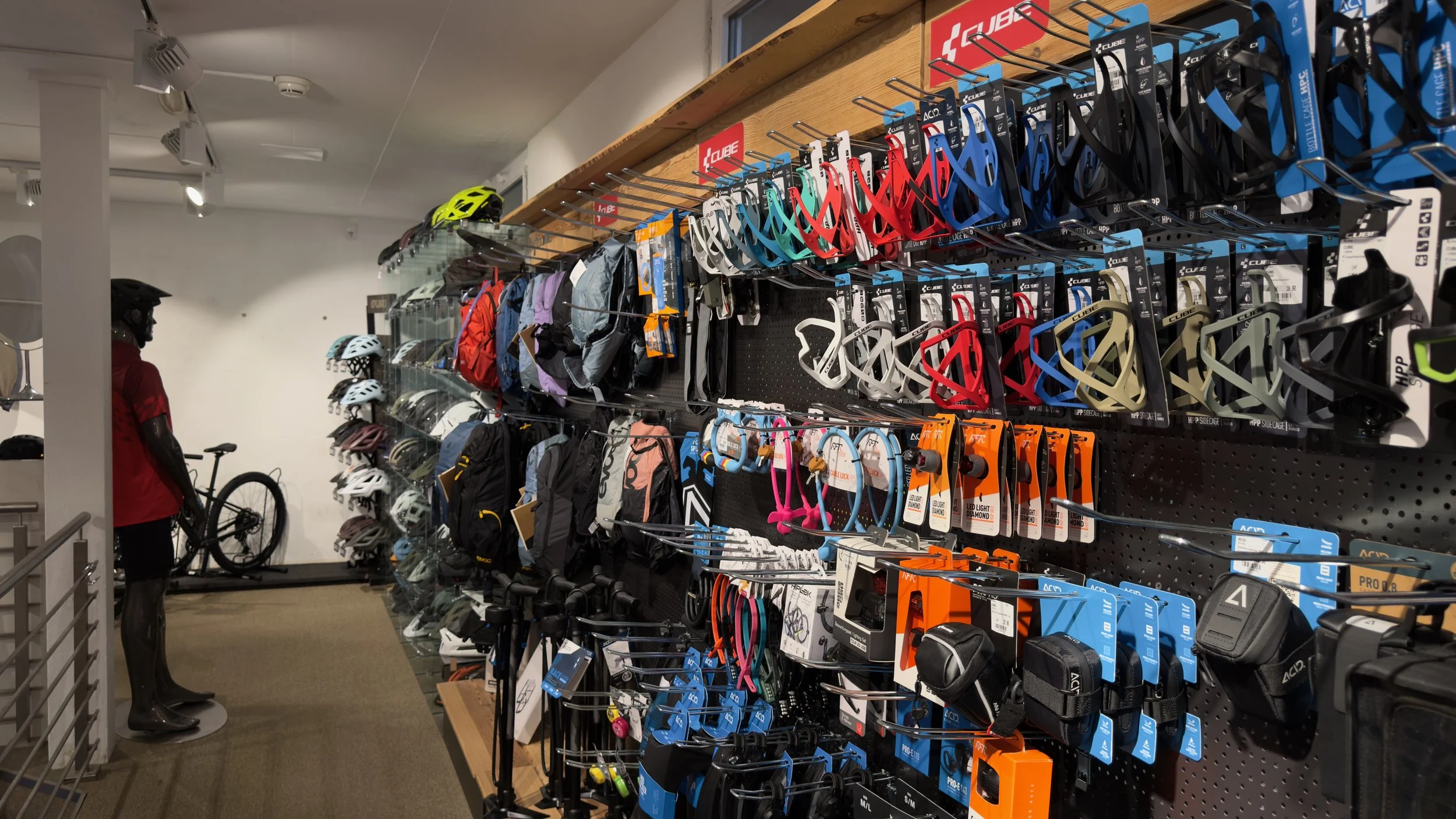 A display shelf in a bicycle shop featuring bicycle accessories, helmets, water bottles, pedals, and sunglasses; in the background, a bicycle and a person wearing a red shirt and a bicycle helmet.