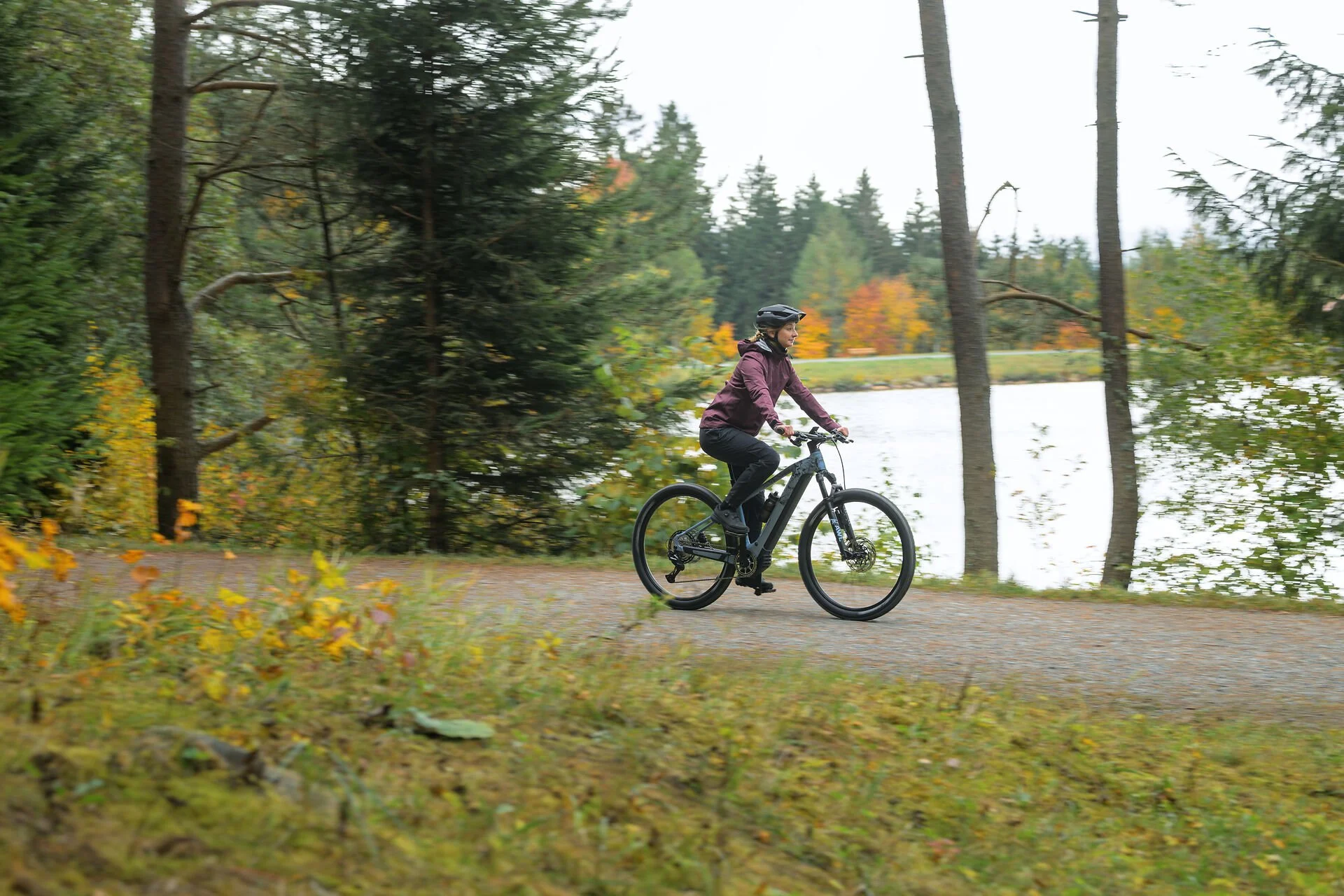 Frau fährt mit einem Mountainbike auf einem Waldweg neben einem See, umgeben von Bäumen im Herbst.