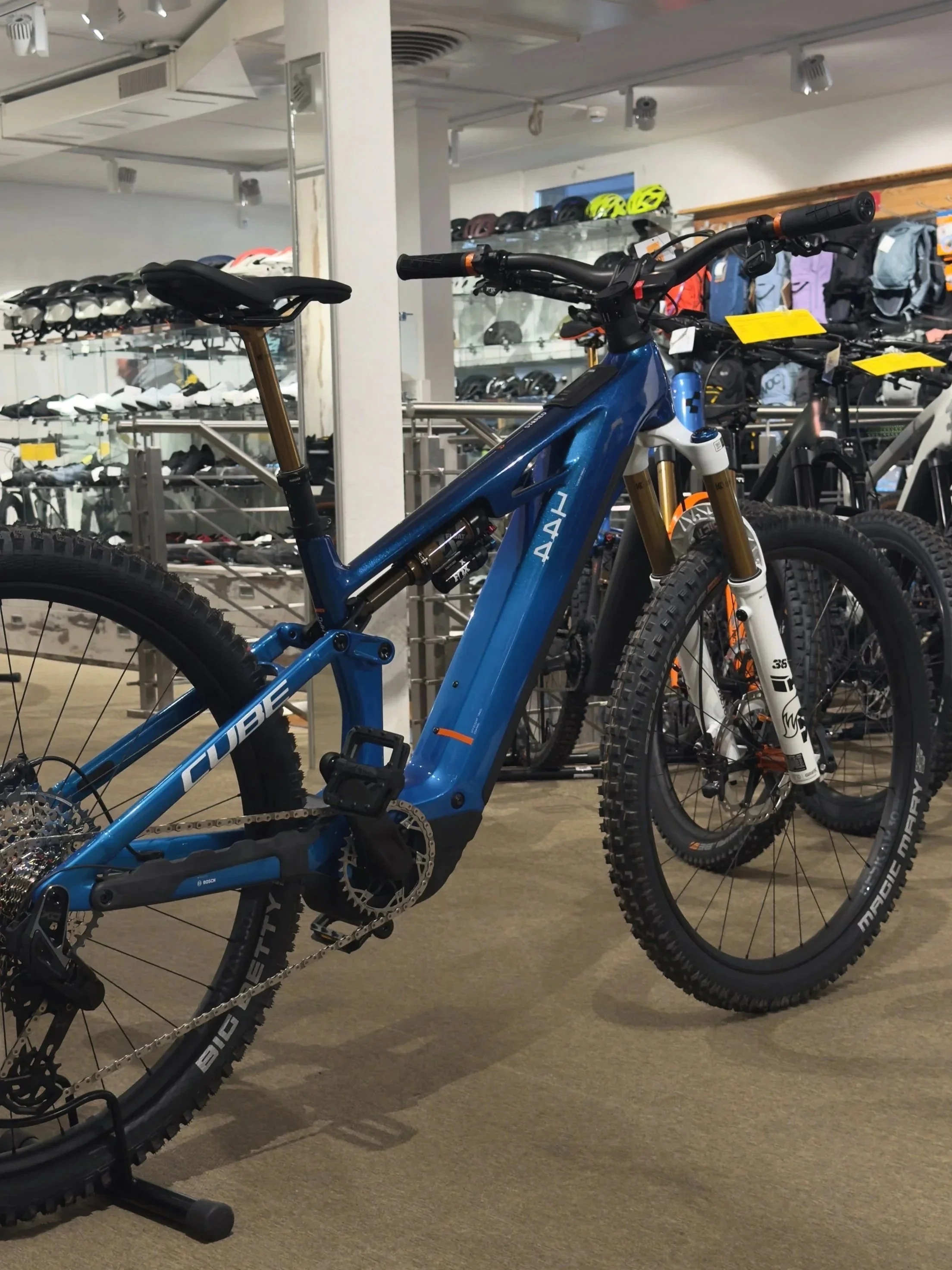 A blue mountain bike in a bike shop; helmets and backpacks can be seen on shelves in the background.