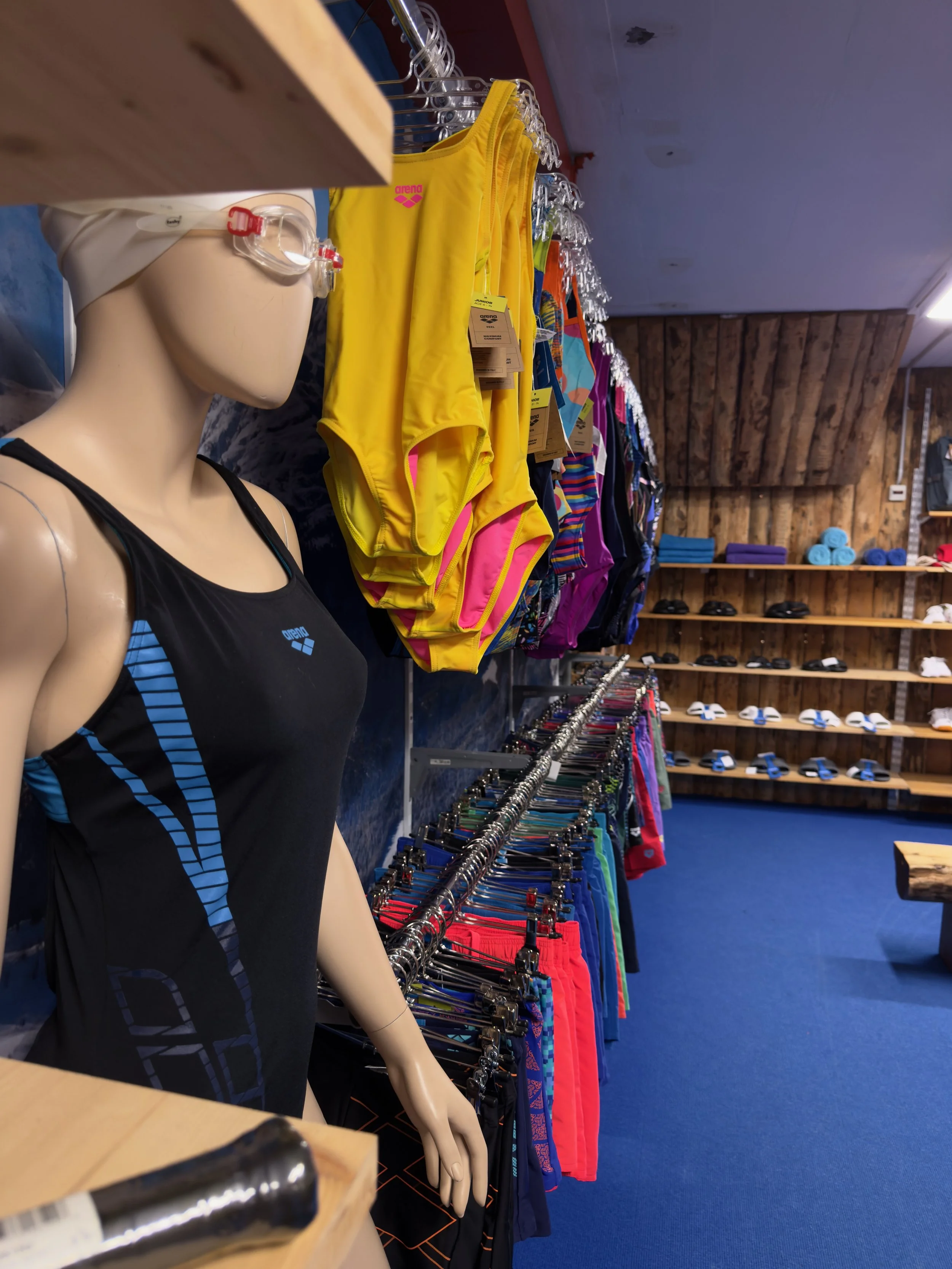 A store selling swimwear and towels, displayed on wooden shelves against a wooden wall. Blue, purple, and red towels are stacked on one shelf. In the foreground is a mannequin wearing a black swimsuit with blue accents and a pair of goggles.