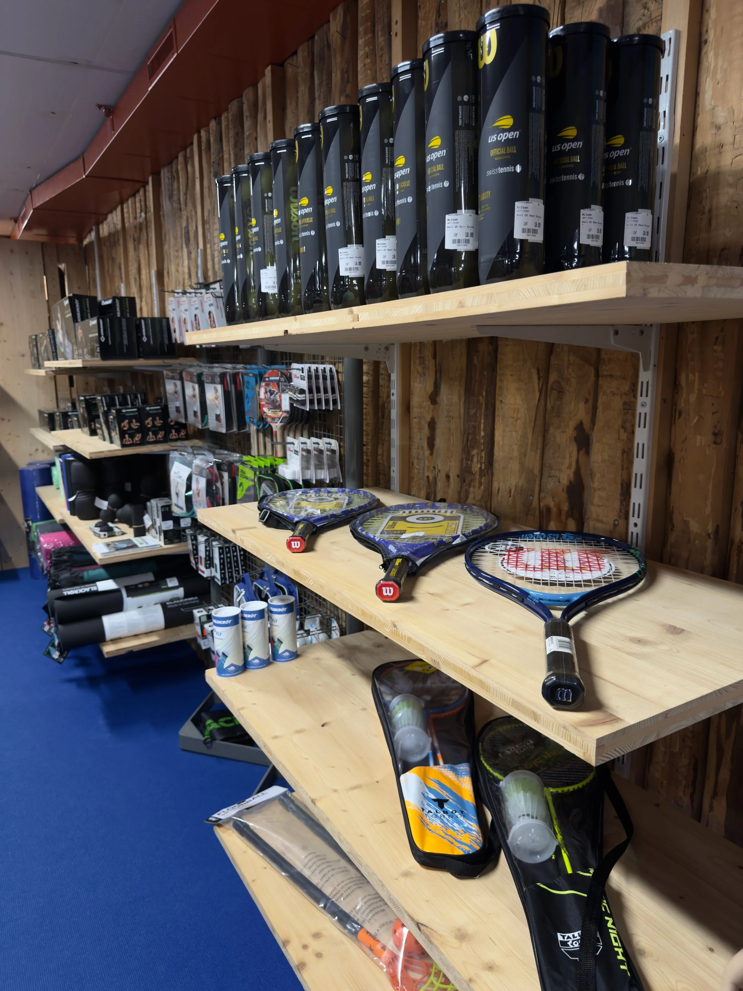 Shelves displaying tennis equipment, including tennis rackets, balls, racket covers, and accessories, in a sports store.