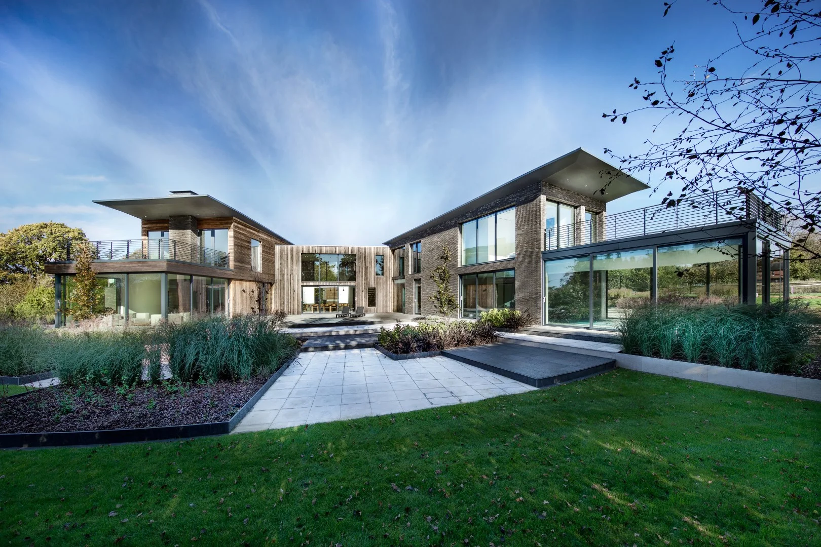The Elements is a private new build house in Hampshire, completed in early 2017 by Winchester based architects AR Design Studio. Adjacent to the stunning River Hamble and its popular marina, it is nestled within meadows rich in native wildlife. With a global collection of friends and family, the client required a large home in which to regularly host and entertain. The home focuses on accommodating a number of guests in the homeliest way possible, providing an experience of a ‘home-from-home’. It is hoped that the home will create lasting memories among visiting friends and family. The massing for the home is divided into three interlocking blocks. Two blocks, one for guests, the other for the family, are connected by a central entrance block. The ground floor becomes a shared zone focused on leisure, socialising and entertainment. The team used the path of the sun to carve out three garden zones, grouping lifestyle activities based on daily rituals. The leisure facilities of the pool, games room, gym and tennis court all receive strong morning light. The sun tracks to the central courtyard by midday, creating a sheltered sun-trap that is accessed by the entire ground floor. By the afternoon the sun has moved to the kitchen, lounge and patio, making this side of the house perfect for summer barbequing. Balconies and picture windows on the first floor extend view onto the wider context creating strong elevated visual connections with the gardens. The shaded north contains the arrival and entrance to the property and is designed to compress your view. A wall guides visitors beneath a large cantilever into an entrance courtyard. From here you enter the house into a double height space that opens up to the view of the central courtyard and is framed by two staircases and walkway. This provides access to the homes main spaces and connects the guest and family blocks. Glazing is extensively used throughout the house to blur the distinction between garden and house and provides connecting views across the open, free-flowing plan and to the gardens beyond. The Elements represents an ambitious taste of shared living, designing not only for the clients but for their family and friends. It has become a perfect reflection of the clients dream; a living, breathing space, seamlessly connected to its surroundings.