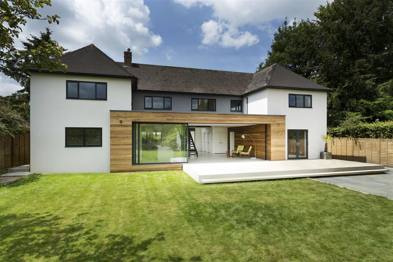 Nestled along a leafy lane on the outskirts of Winchester sits Kilham House. Once a tired building with a confusing layout, the house now boasts a contemporary update that really transforms the house into the five-bedroomed family home that it desperately needed to be. A grand, double-height entrance guides you into the building, immediately bringing you into the heart of the home which has now been become the main living space for the family. A large expanse of sliding glazing gives views into the garden, allowing the three children to run wild whilst the parents can relax in the central space and still keep a watchful eye. A key and exciting feature of the house is the staircase. Centred in the property it acts as a locus to the project, dividing the space between the kitchen, dining area and the living areas. Steel wires hang around the staircase, enclosing it in a contemporary wrap whilst also forming part of the balustrade. The stairs take you up to the first floor and onto a bridge that flows across the double height entrance space. A tongue in cheek use of Foscarini’s Gregg pendant lights give a feeling of being up in the clouds, adding to airy and spacious feel of the central space. At the rear of the property a central timber form connects the two wings of the house and projects into the garden creating an architectural form that ties the whole project together. A large concrete plinth that steps down to the garden creates a place to relax and dine outdoors. The concrete plinth flows into the property and makes up the entire ground floor surface. This use of material, mixed with the large sliding glazed panels that face onto the garden, blurs the boundary between indoors and outdoors.